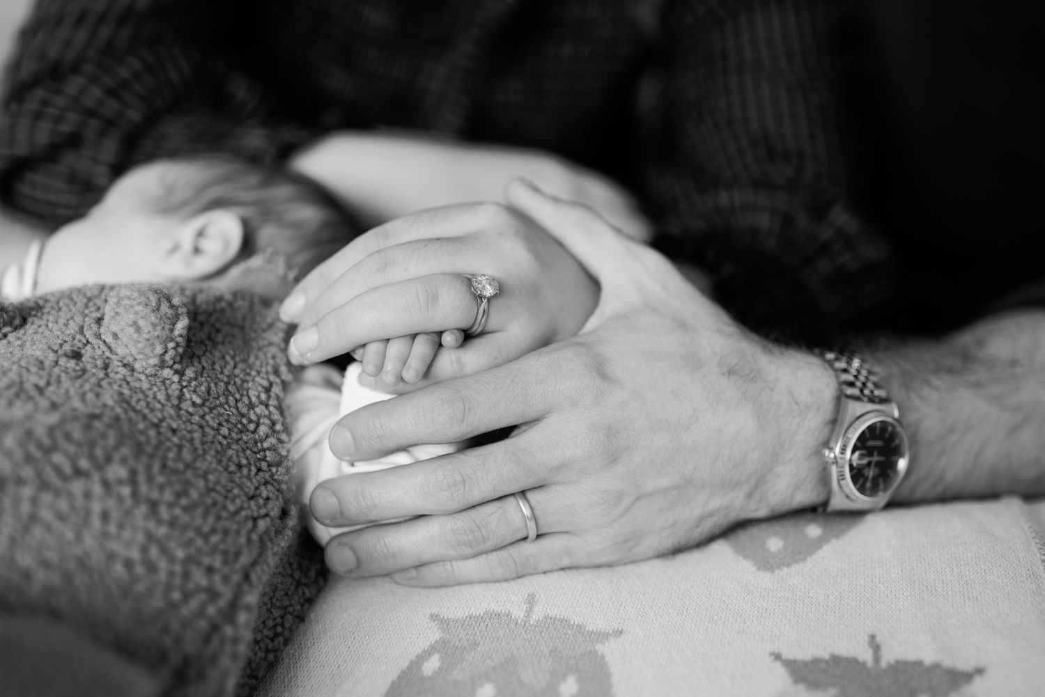 Black and white close-up of a parent's hands, wearing a wedding ring and watch, gently holding a sleeping newborn baby's tiny hand.