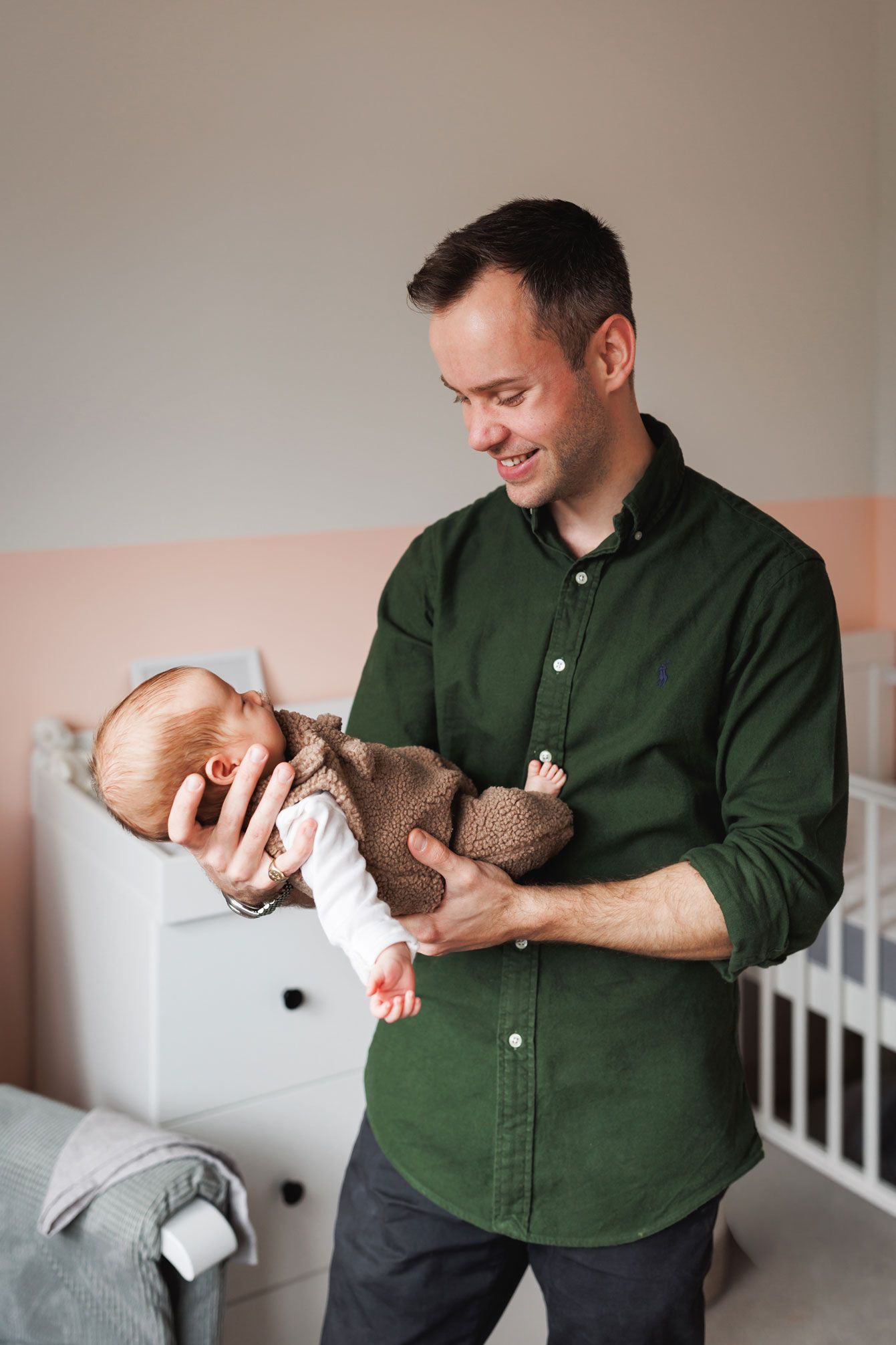 Smiling man in a green button-up shirt cradles a newborn baby in a knit outfit in a cozy nursery with a white dresser and crib.