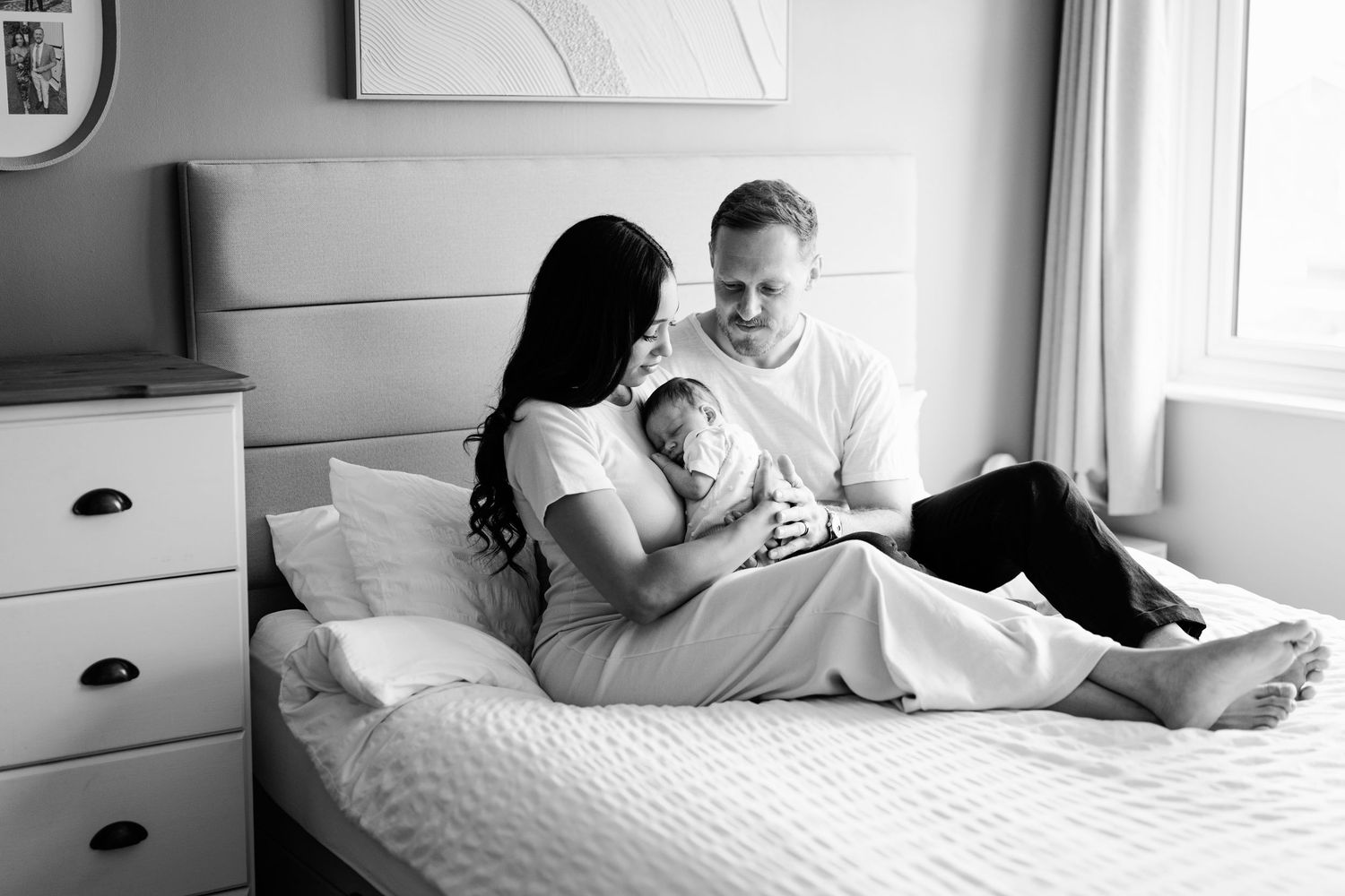 Black and white photo of parents on bed lovingly cradling their newborn baby in a bright bedroom.