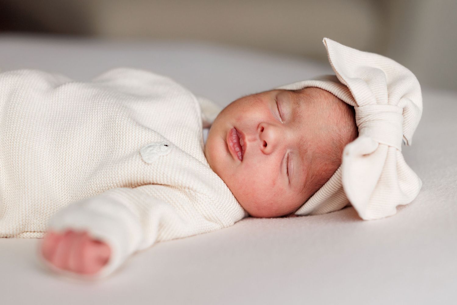 Sleeping newborn baby in a cream knit outfit and matching bow headband resting peacefully on a soft white blanket.