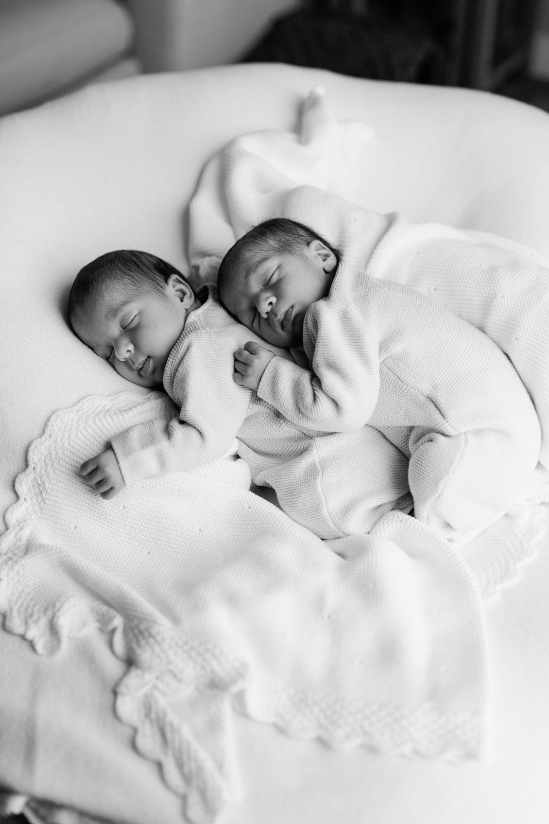 Black and white photo of two sleeping newborn twins in white outfits cuddled together on a soft blanket with delicate lace trim.