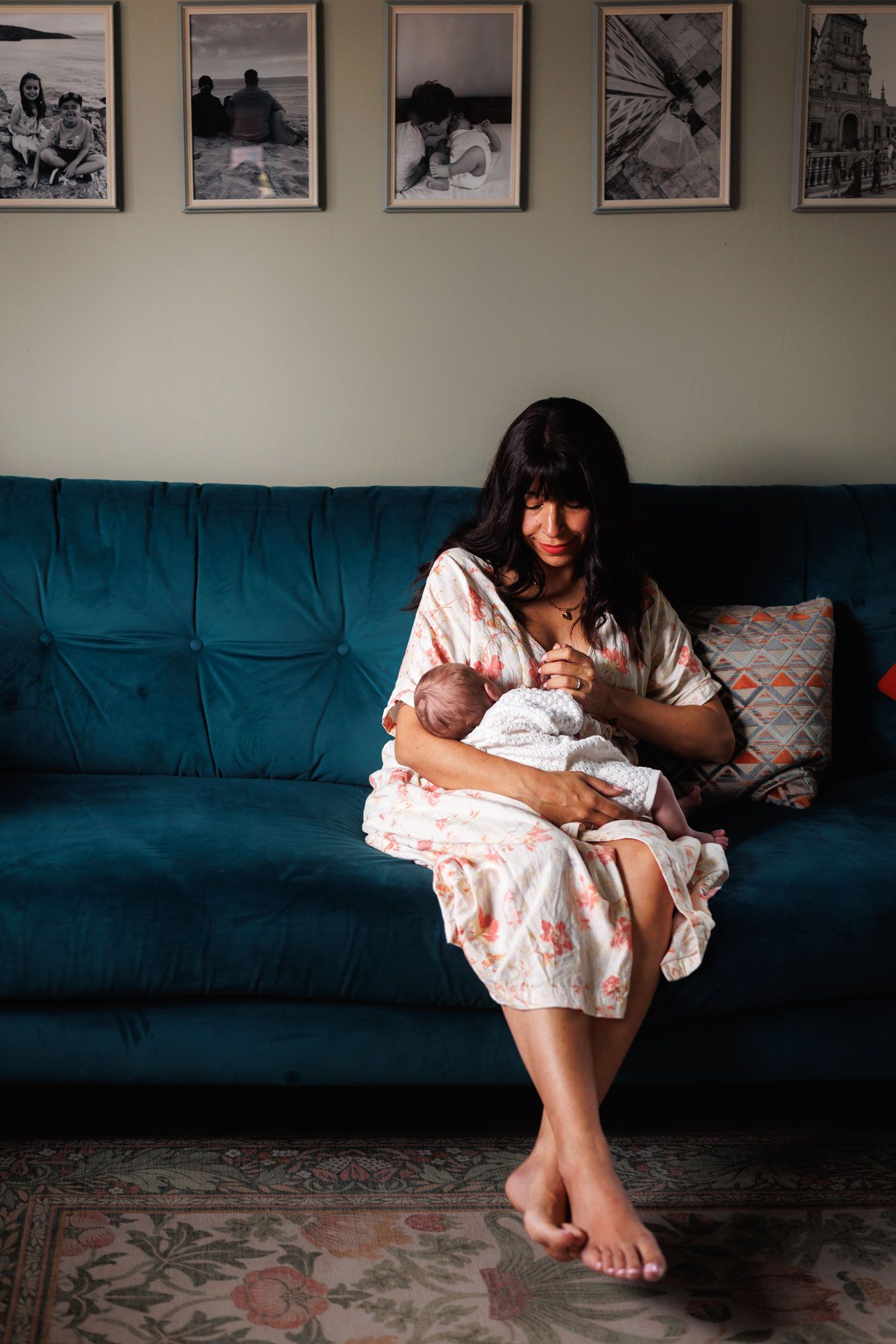 Mother in floral dress tenderly holding her newborn baby while sitting on a teal sofa.