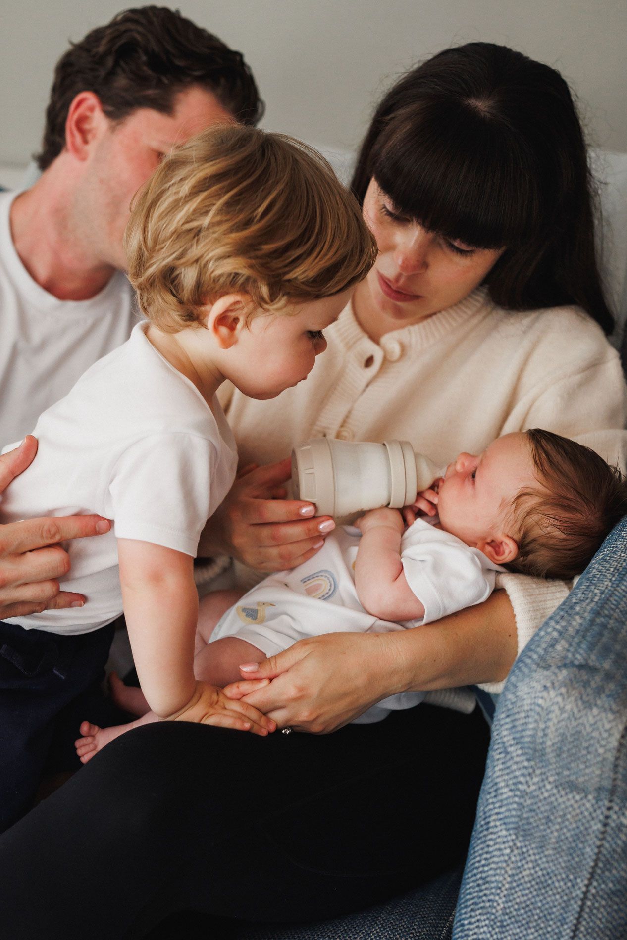 Family of four with mother bottle-feeding newborn baby while toddler watches and father sits behind them.