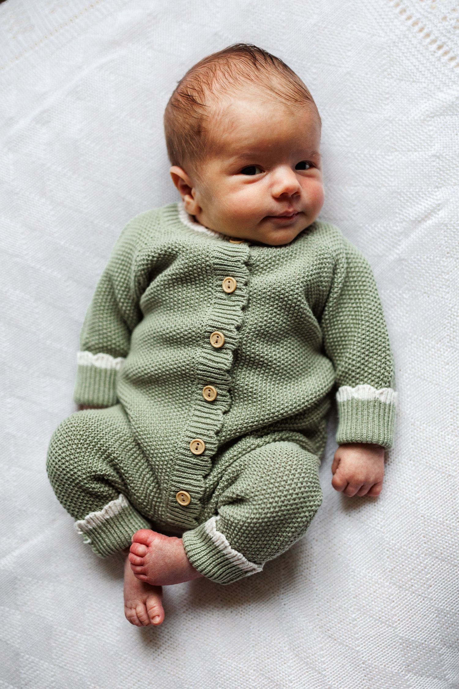 A newborn baby in a green knitted button-up romper lies on a white blanket looking upward.