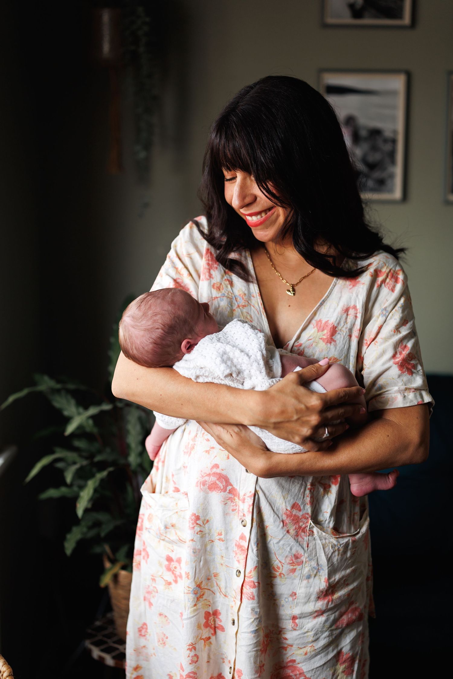 A smiling dark-haired mother in a floral dress lovingly holds her sleeping newborn baby indoors.