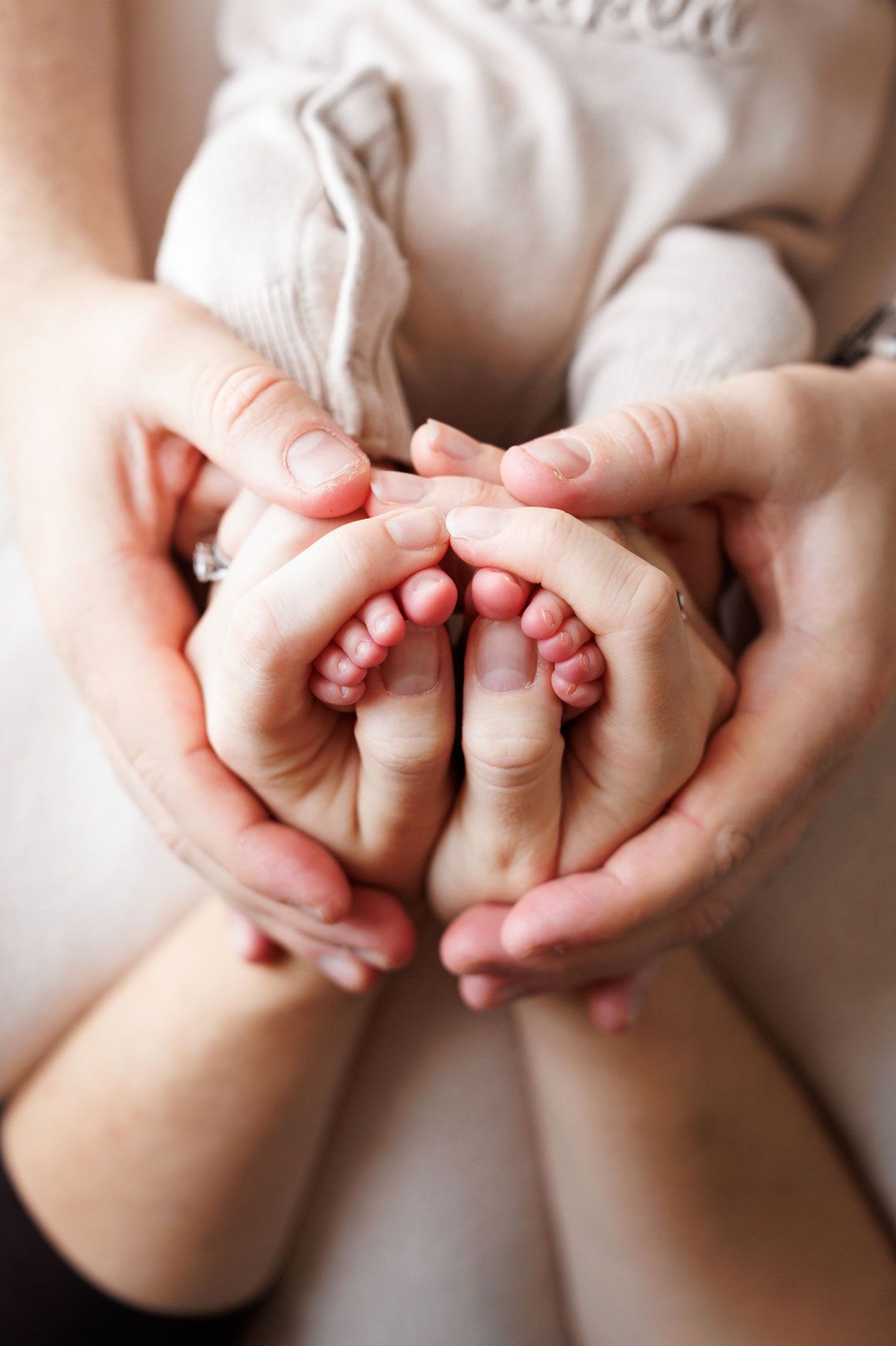 Parents' hands gently cradle a newborn baby's tiny feet against a soft cream blanket in a tender family moment.