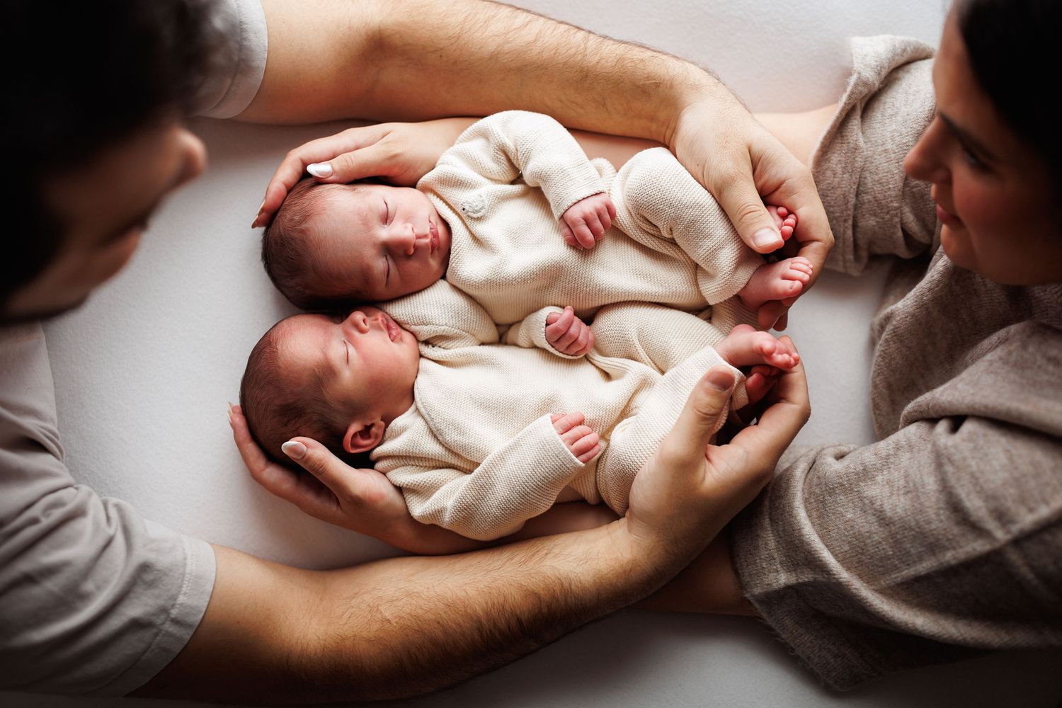 Two sleeping newborn twins in cream outfits lie side by side, cradled in their parents' hands in a soft overhead newborn photo.