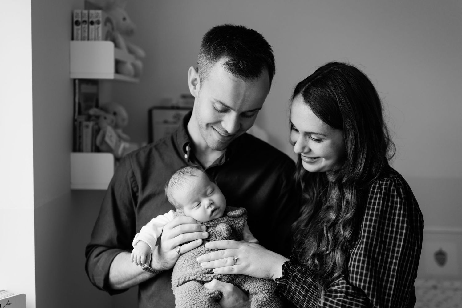 Black and white portrait of smiling parents holding their sleeping newborn baby together in a cozy nursery with shelves behind them.