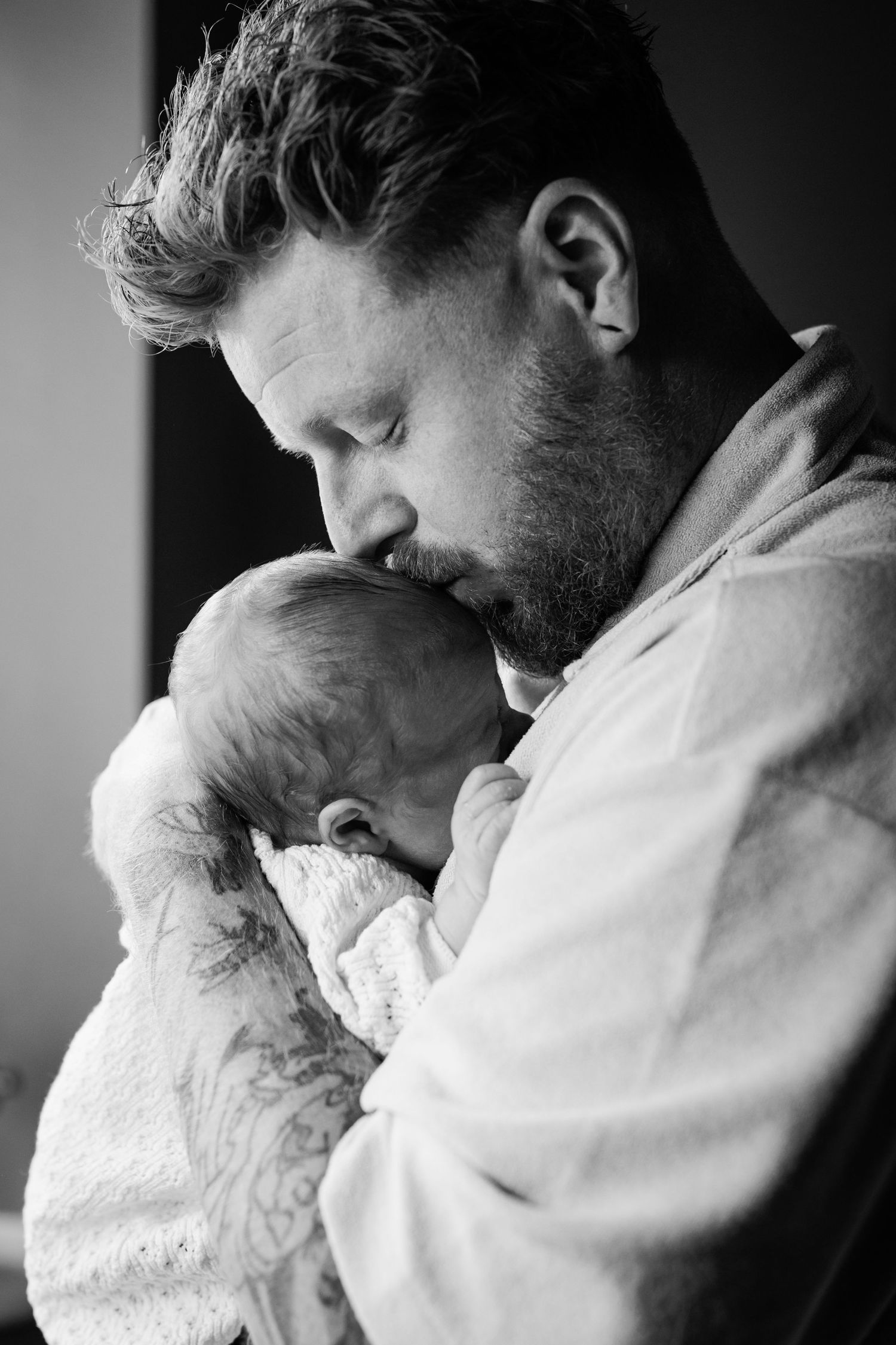Black and white photo of bearded father kissing his sleeping newborn baby tenderly.