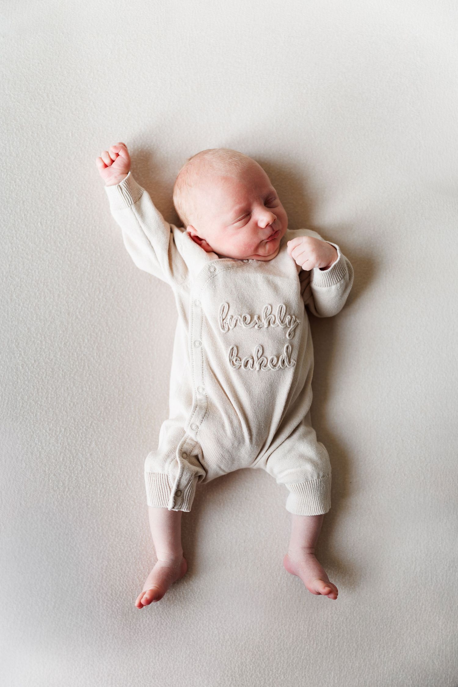 Sleeping newborn baby in a cream knit romper embroidered with the words freshly baked, lying on a soft neutral blanket with one arm raised.
