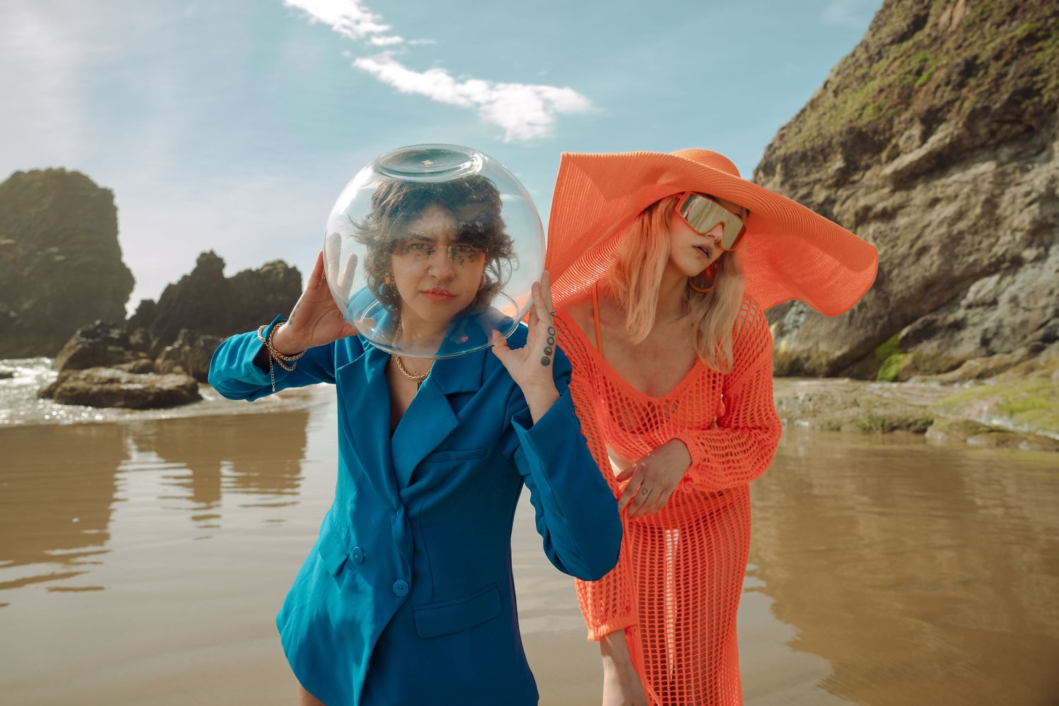Models in bright blue and coral outfits pose playfully on beach with dramatic coastal cliffs.