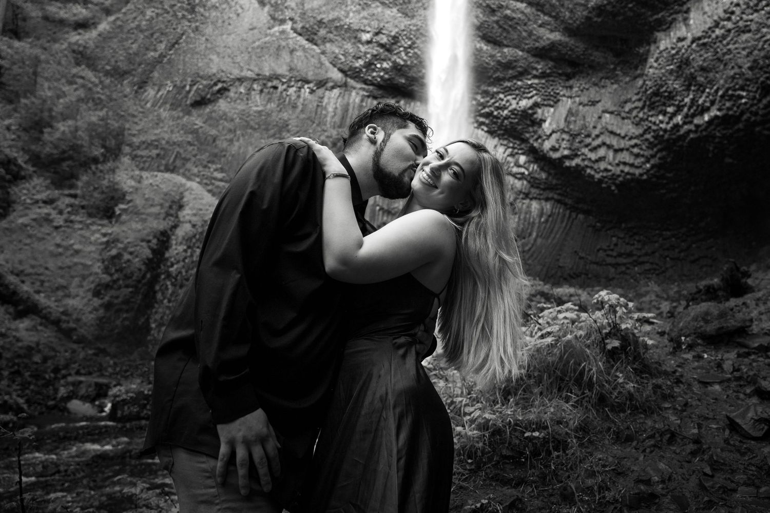 Romantic couple kissing beneath a majestic waterfall in black and white.
