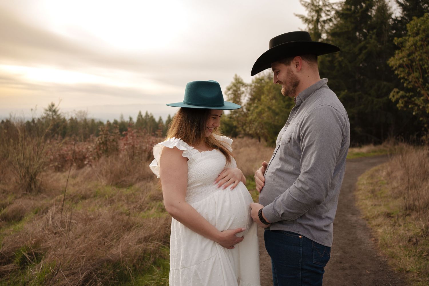 Expectant couple in western wear sharing tender moment in golden field at sunset.