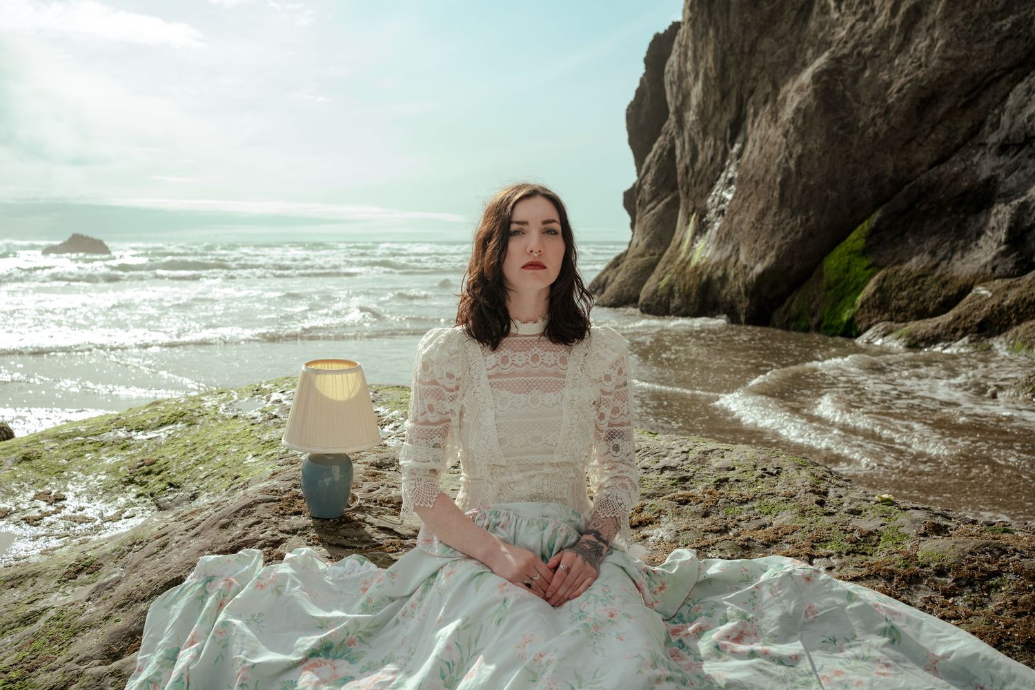 Dreamy coastal scene with lace dress and blanket on rocky beach during overcast day.