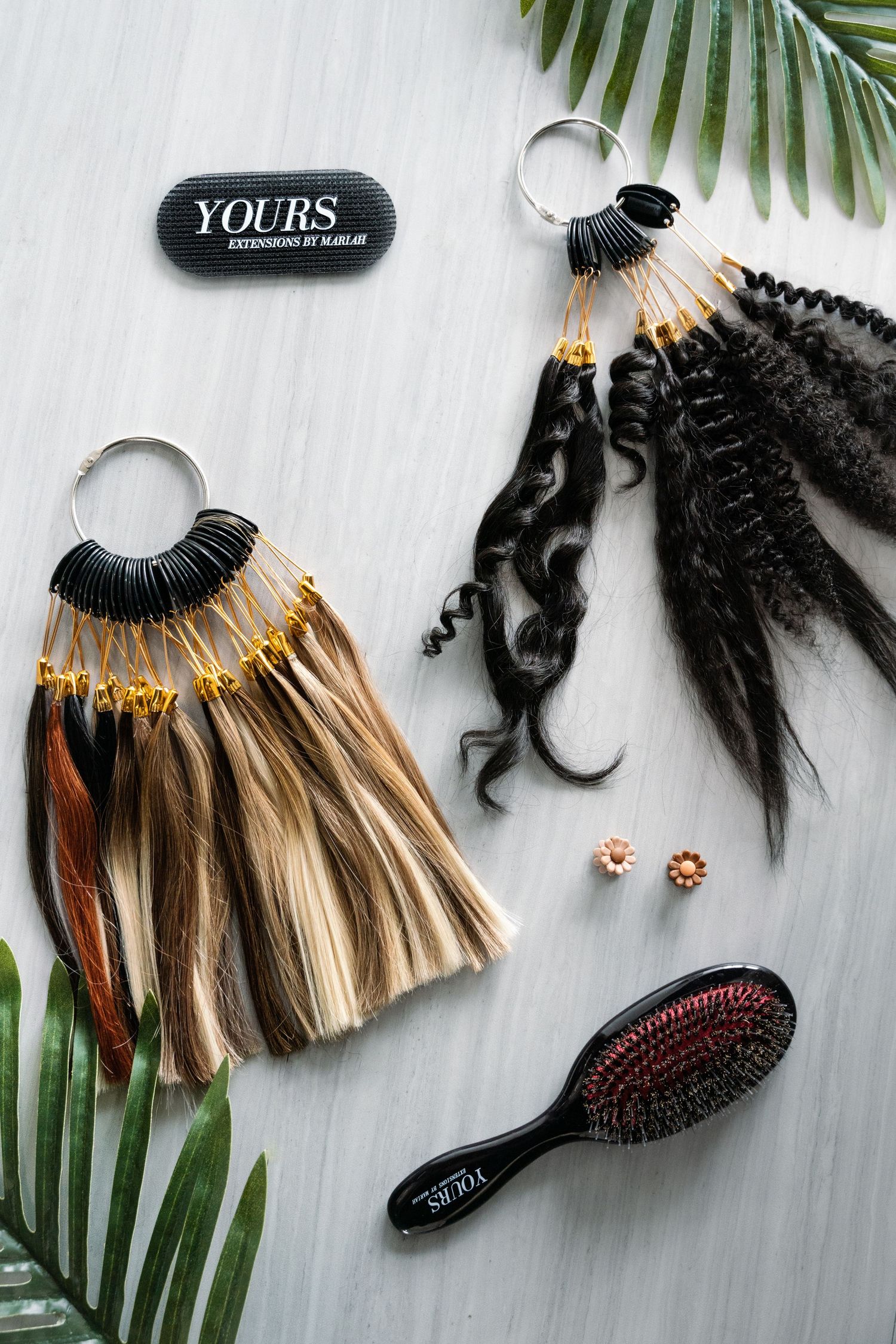 Hair extension samples and styling tools arranged with palm leaves on white background.