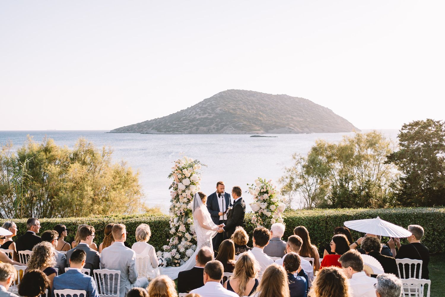 Outdoor wedding ceremony with guests seated overlooking a serene sea and island backdrop under clear blue skies.