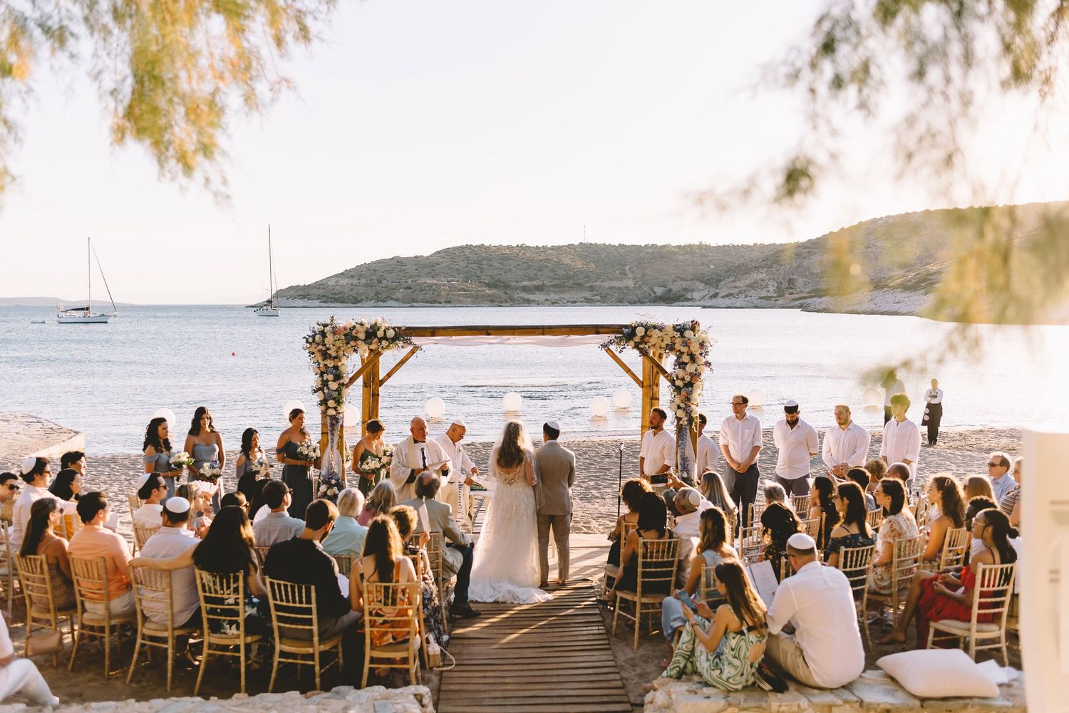 Outdoor beach wedding ceremony with guests seated on wooden deck, floral arch, and sailboat in background at sunset.