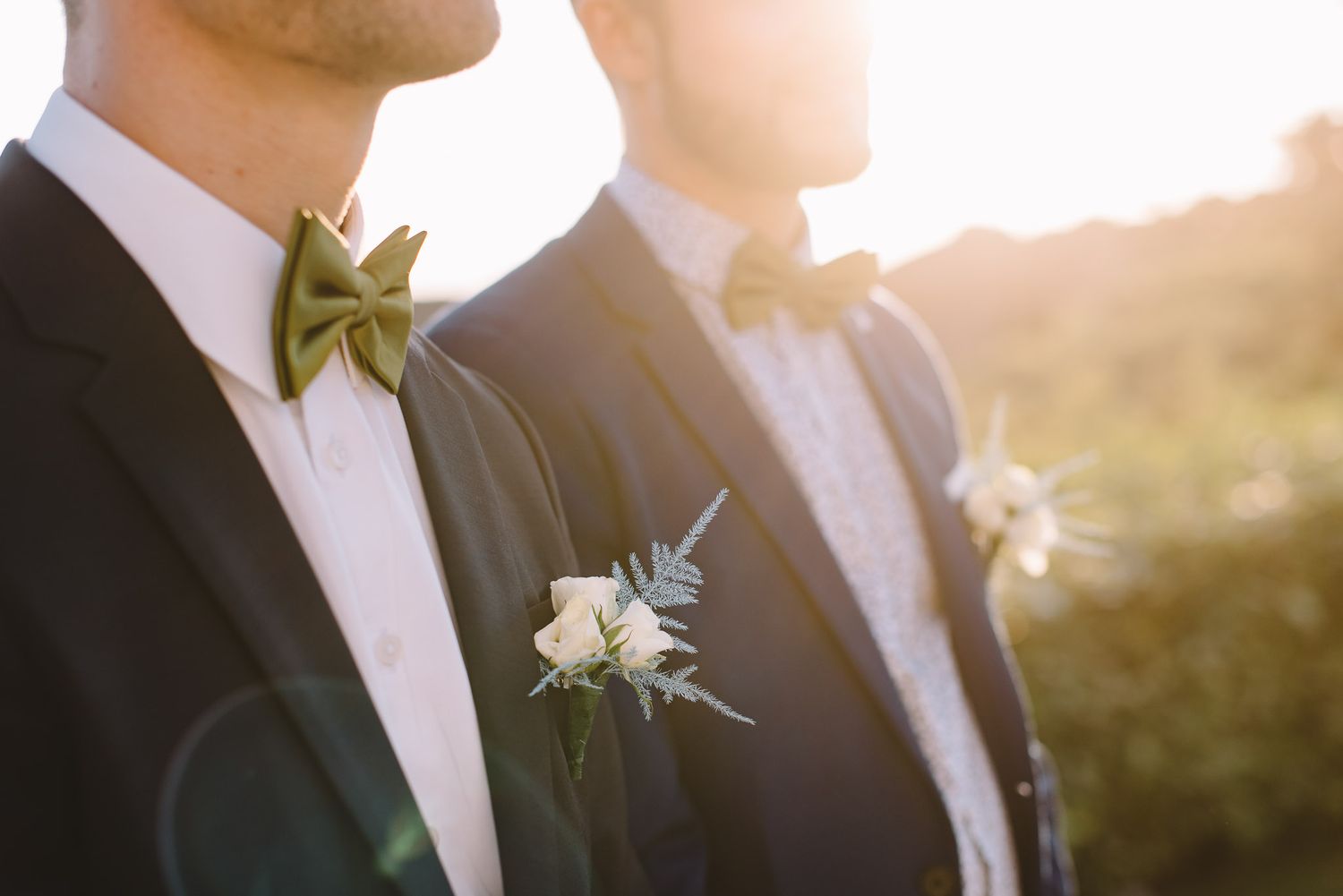 Two grooms in navy suits with olive green bow ties and white boutonnieres stand together in golden sunset light.