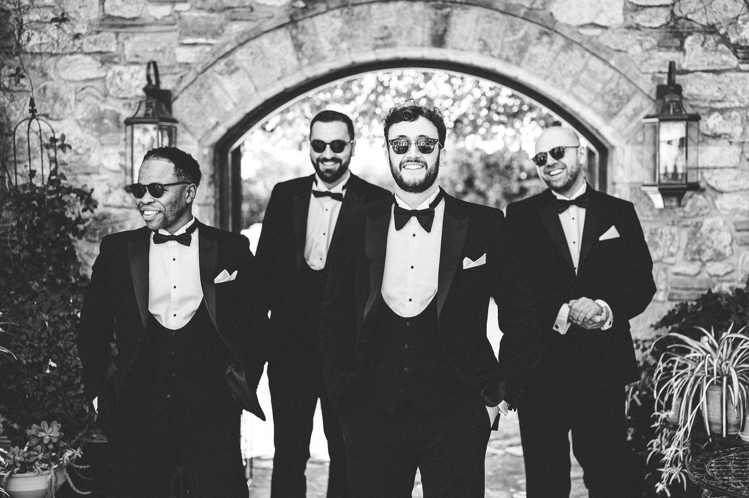 Four groomsmen in black tuxedos and sunglasses pose confidently before a stone archway in this elegant black and white photo.