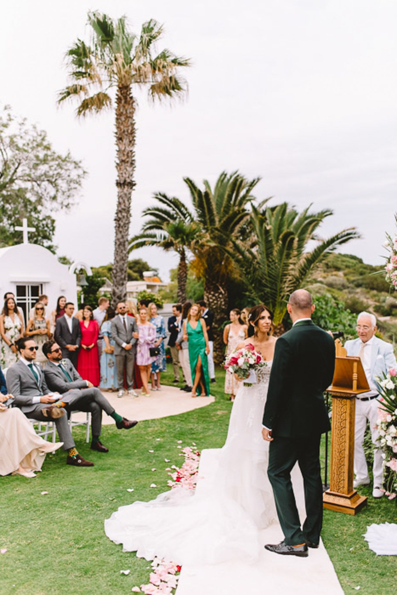 Outdoor wedding ceremony on green lawn with bride and groom at altar, guests seated, palm trees and white chapel in background.