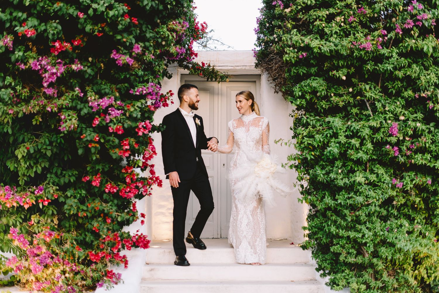 Bride and groom walk hand-in-hand through a floral archway of bougainvillea and ivy at an elegant outdoor wedding.