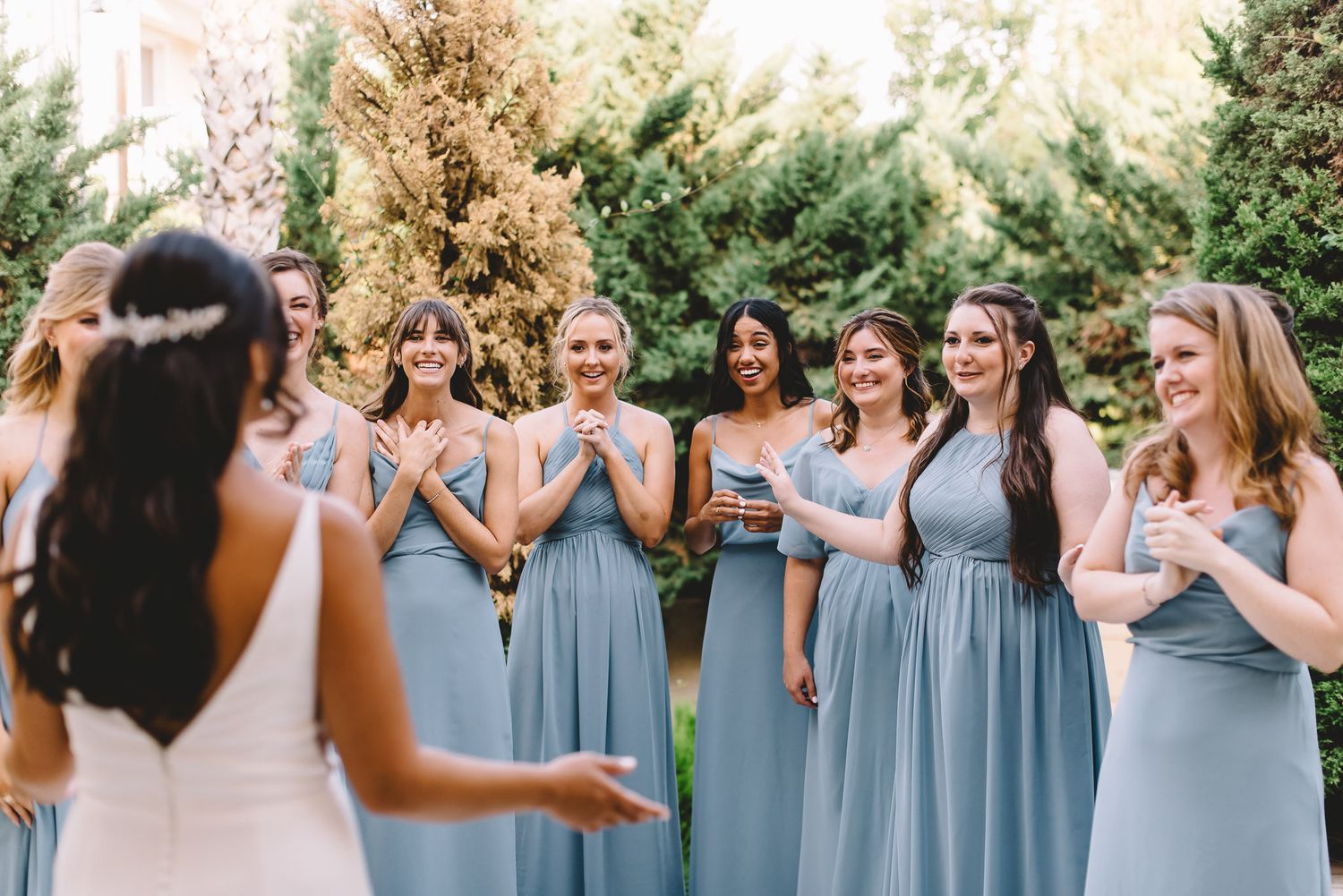 Bridesmaids in dusty blue dresses react with joy as bride reveals her wedding gown in an outdoor garden setting.