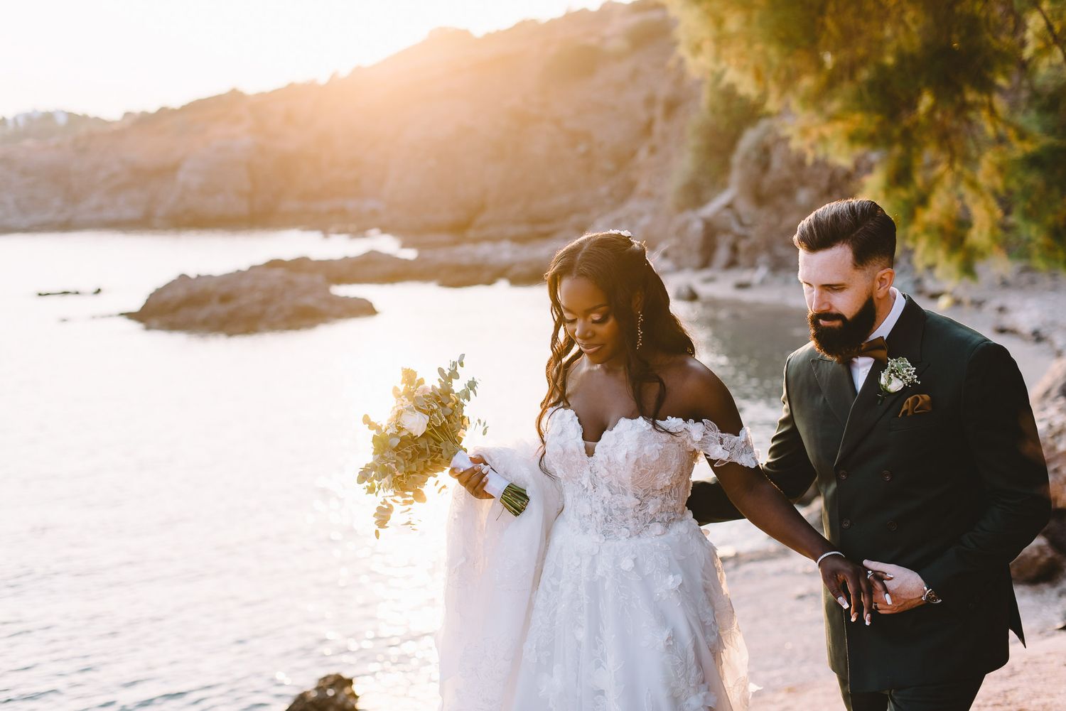 Bride and groom walk along rocky coastal shore at golden sunset, bride holding floral bouquet in elegant white gown.