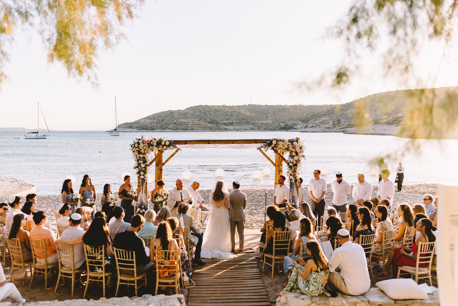 Romantic beach wedding ceremony at sunset with guests seated on wooden deck overlooking calm sea and hills.