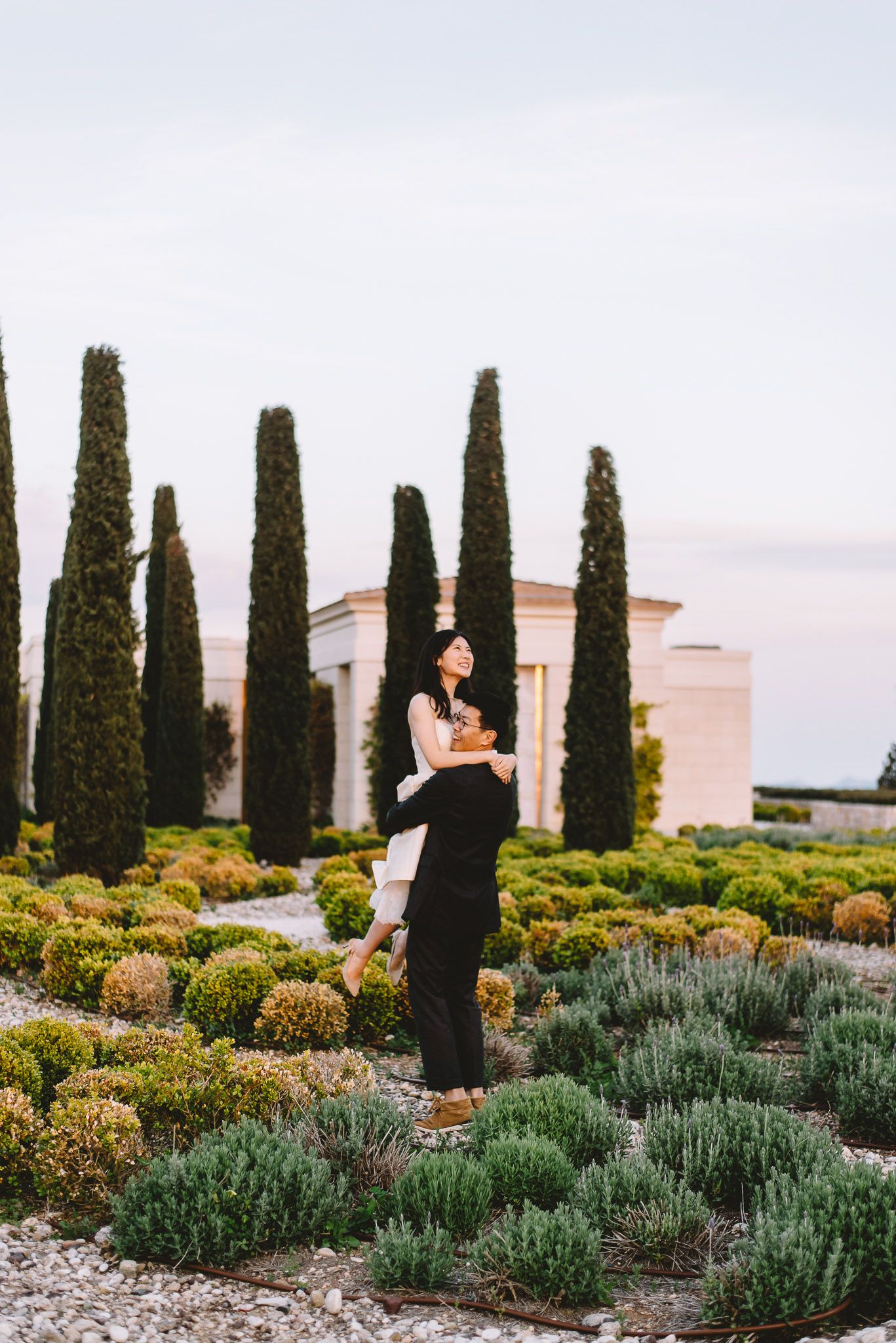 Couple embracing in a formal garden with cypress trees and manicured hedges at dusk.