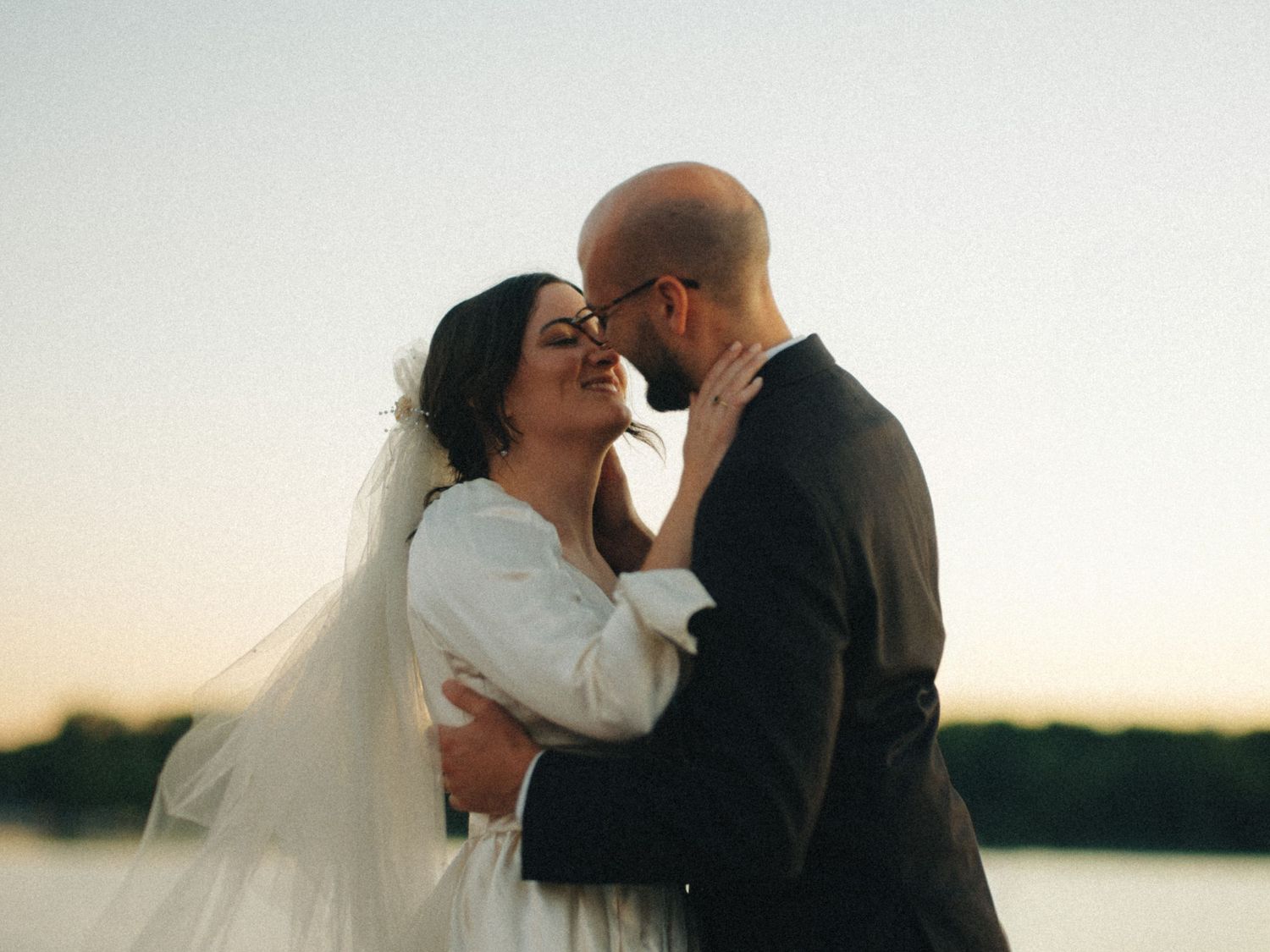 A romantic sunset moment between a couple in wedding attire near a waterfront.