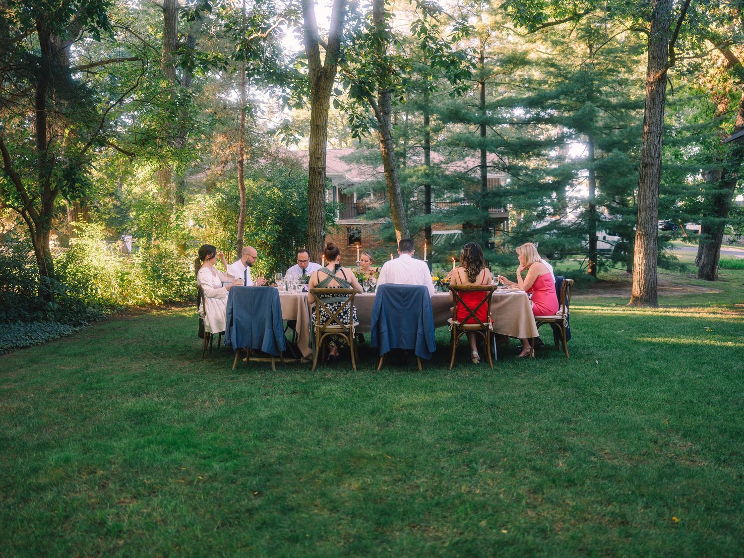 Group of people dining at a long table under trees in a backyard garden setting.