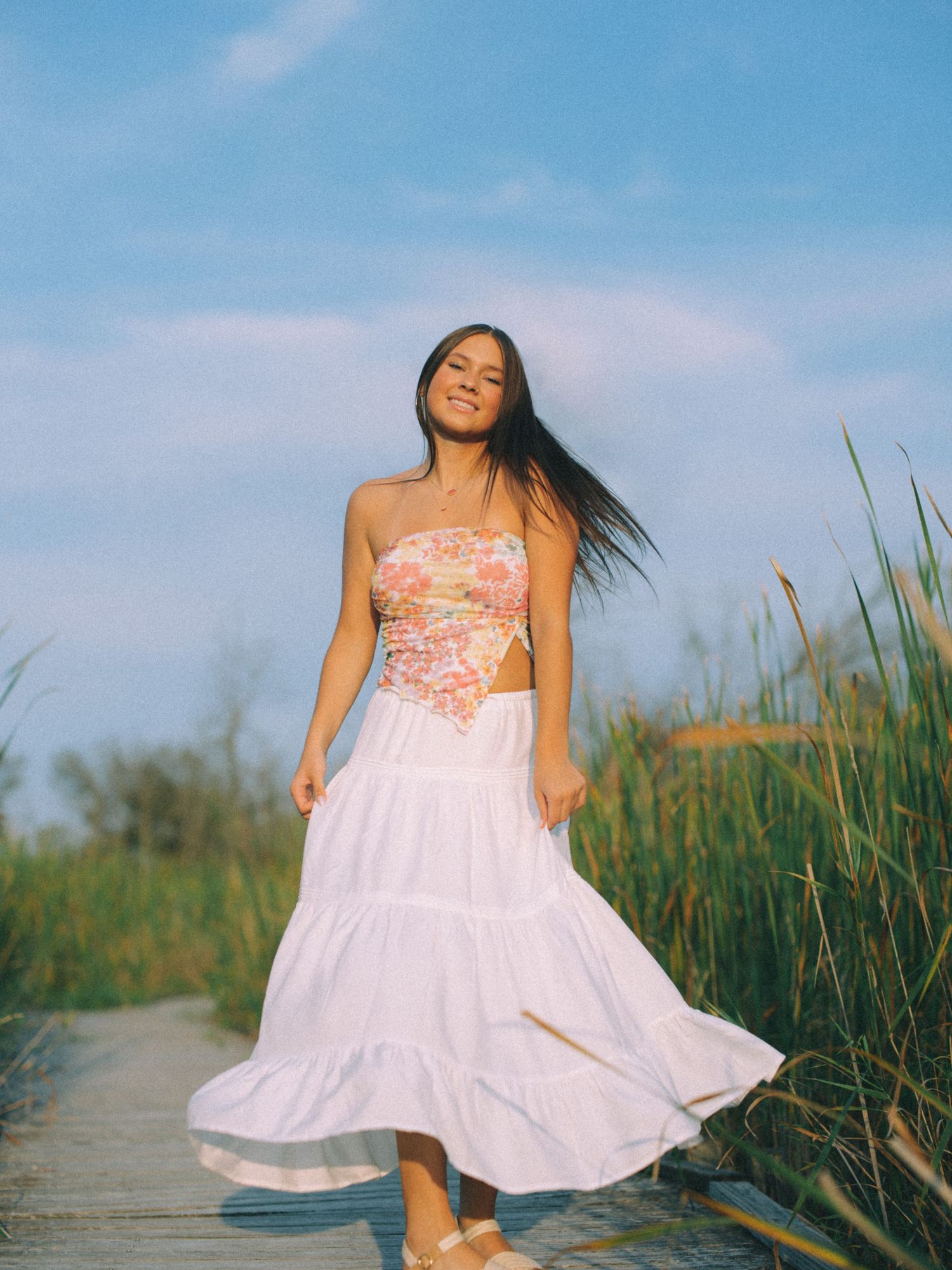 Person in summer dress posing on boardwalk with flowing fabric and natural backdrop.