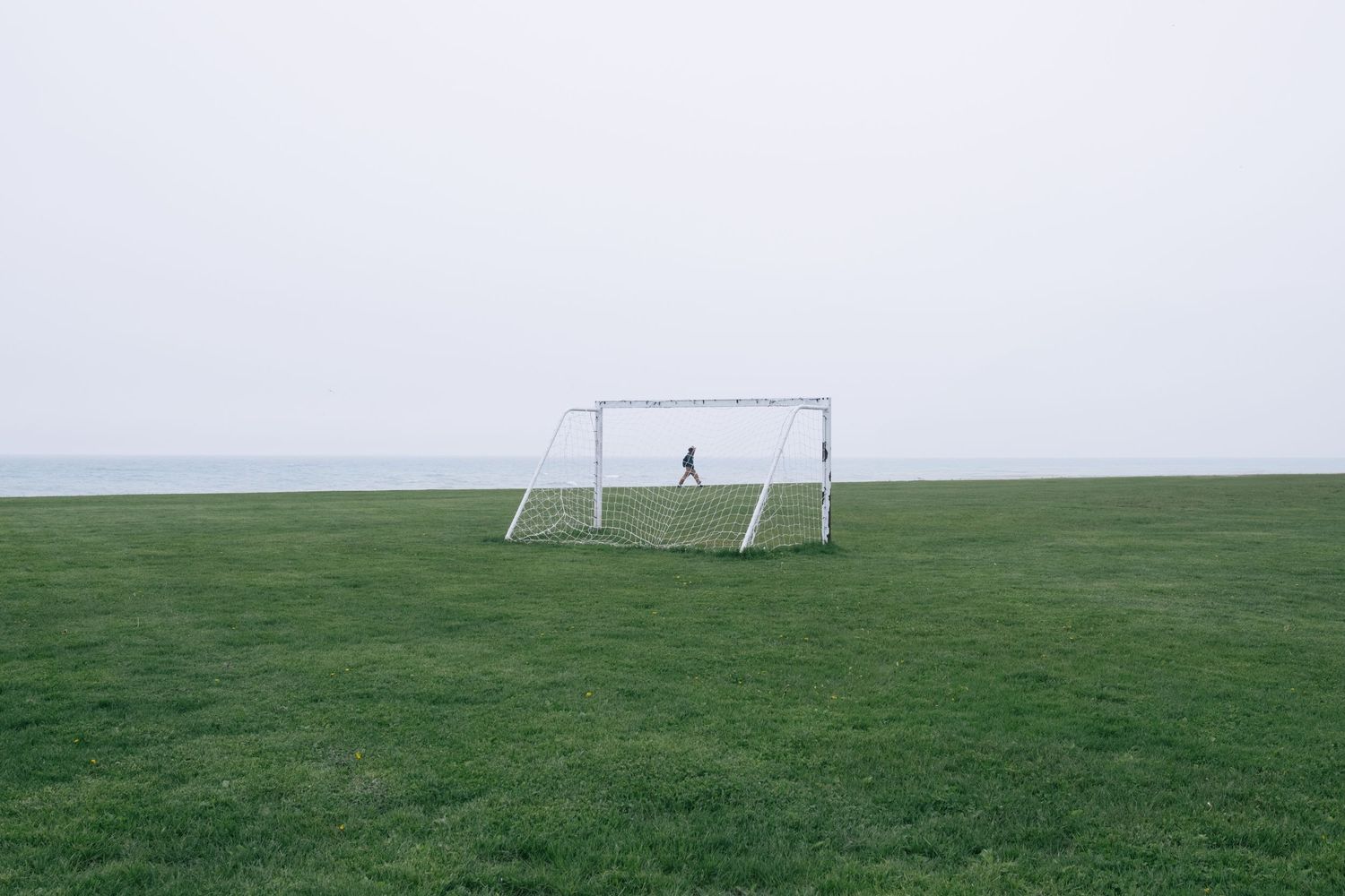 A solitary soccer goal stands on an empty grass field against an overcast sky.
