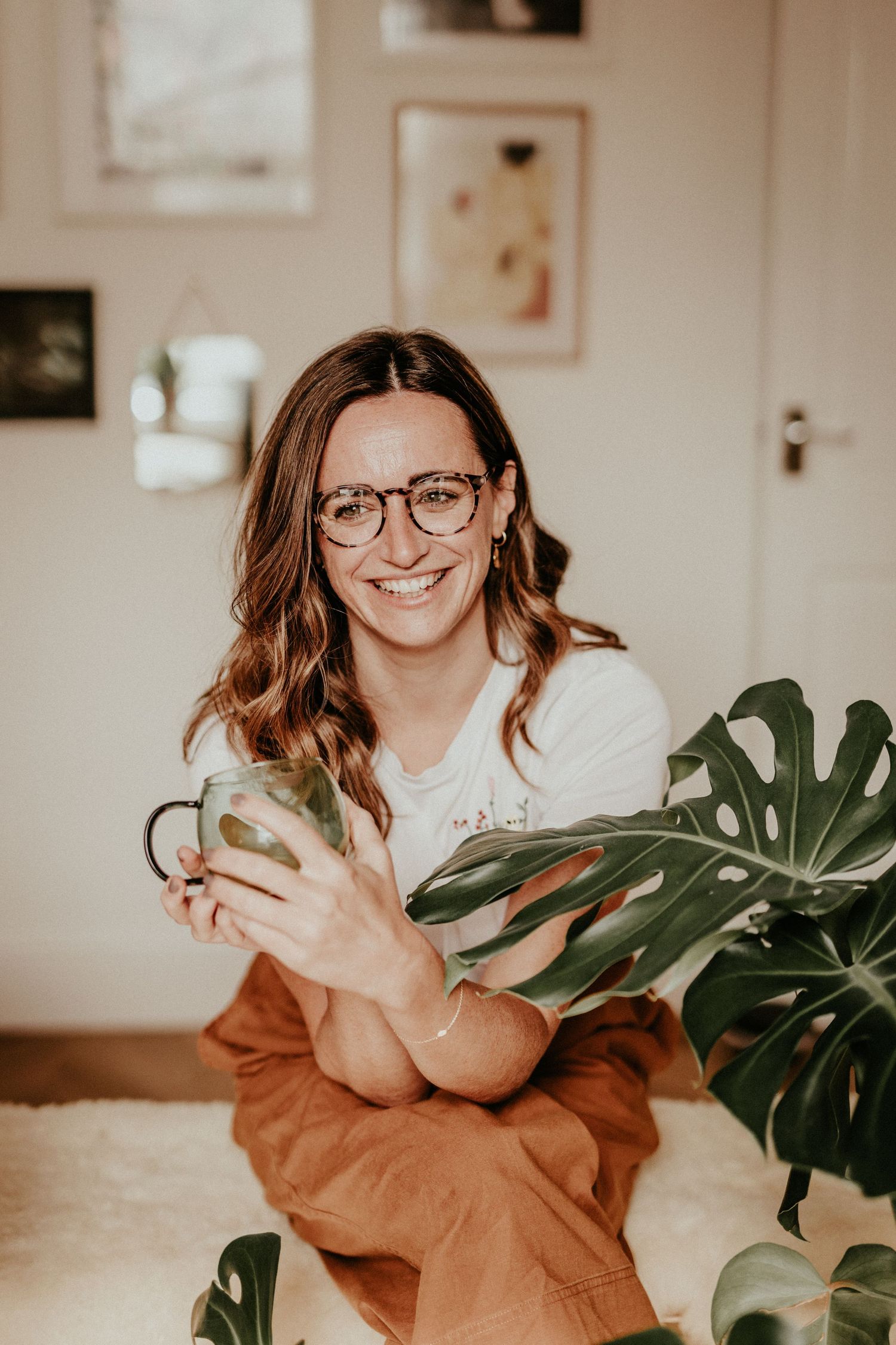 A casual indoor portrait with monstera plants showing someone wearing glasses and a rust-colored cardigan.