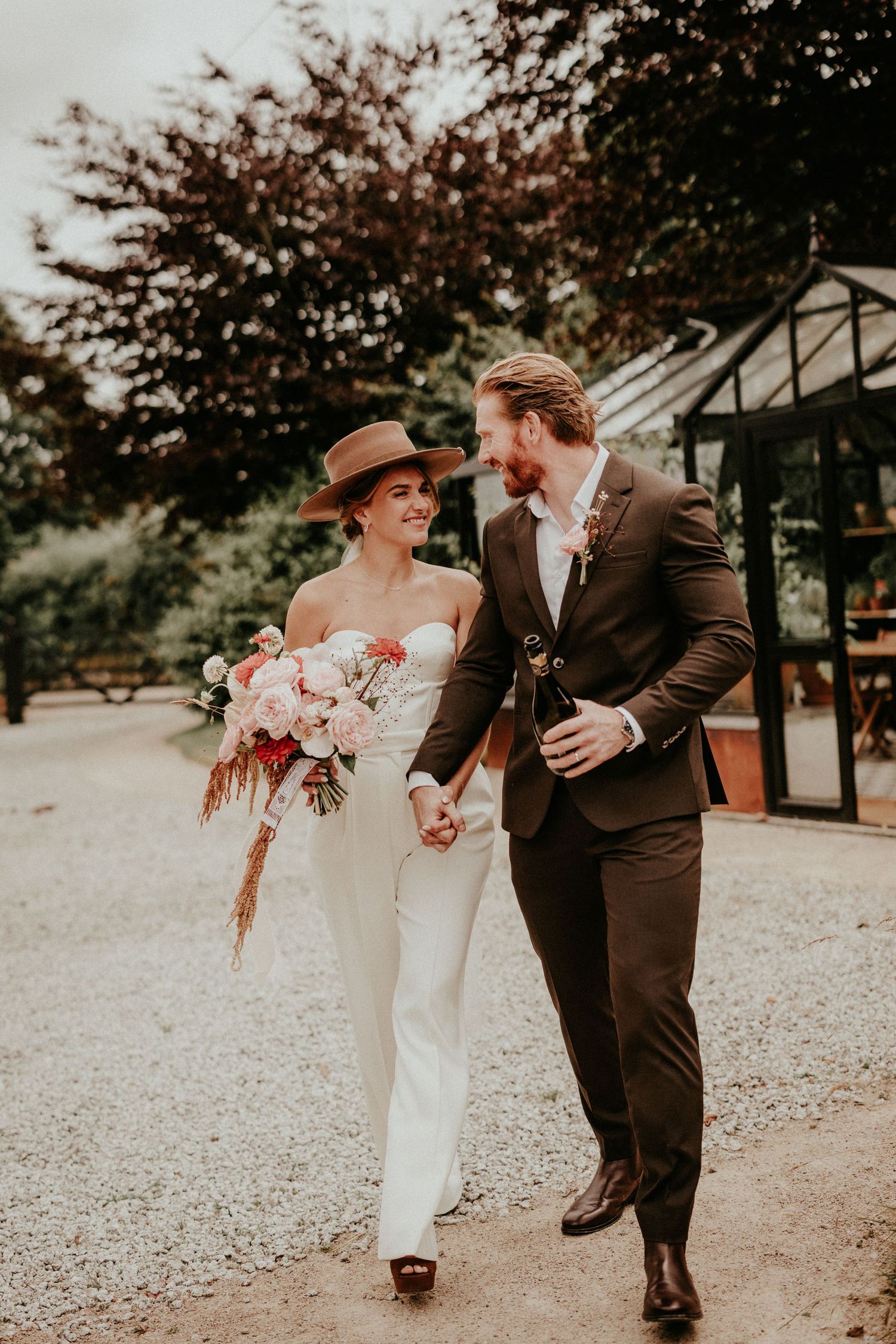 Elegant couple shares candid laughter while walking through gravel path with pink floral bouquet and brown attire.