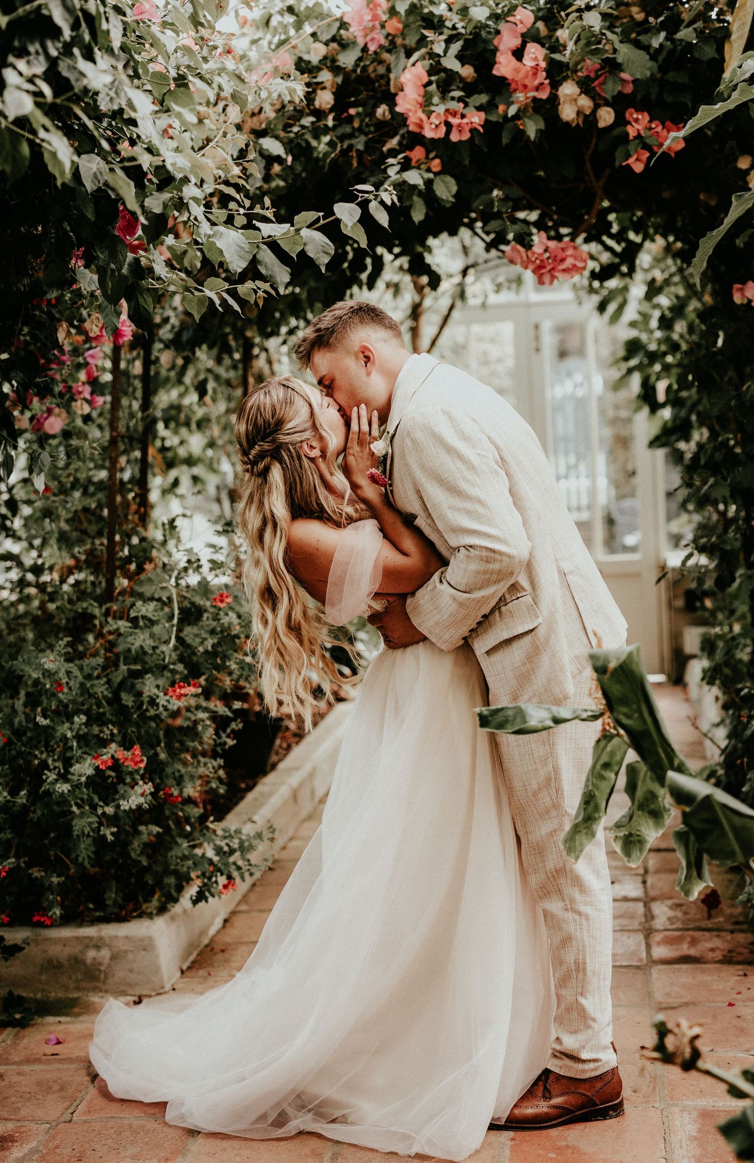 Wedding couple shares a romantic kiss under blooming pink flowers in a garden archway.
