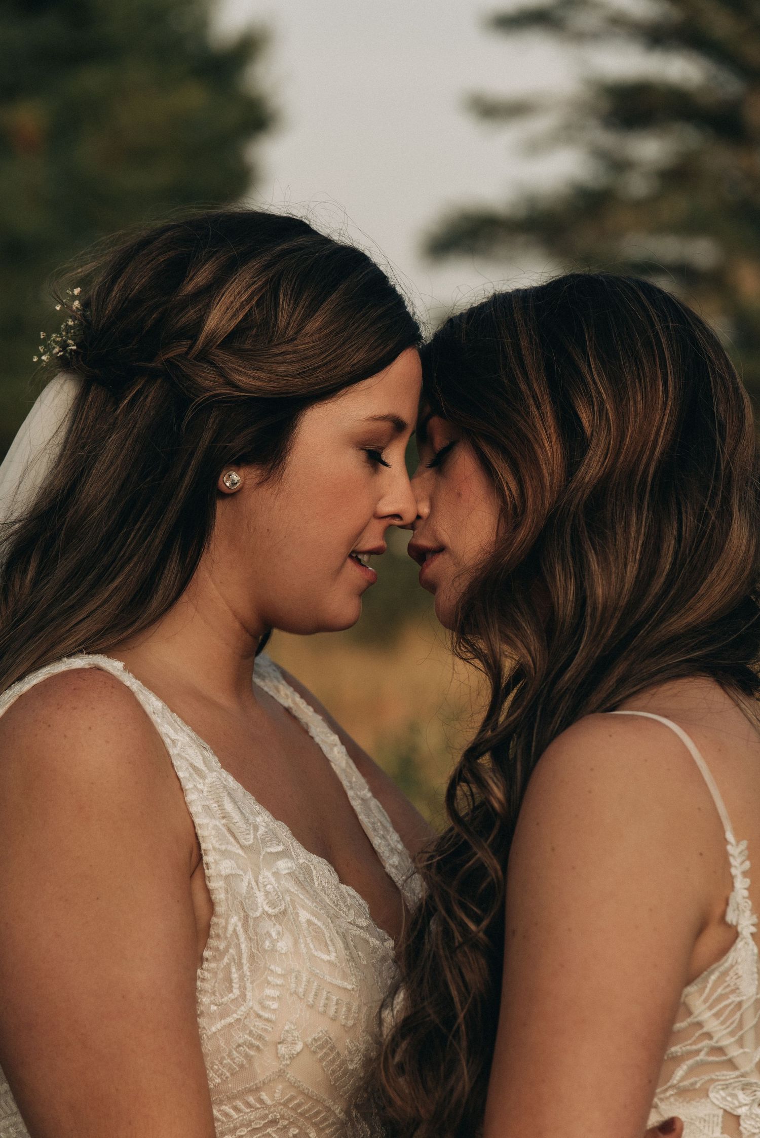 Two people in white lace wedding dresses share an intimate moment with foreheads touching during outdoor photo session.