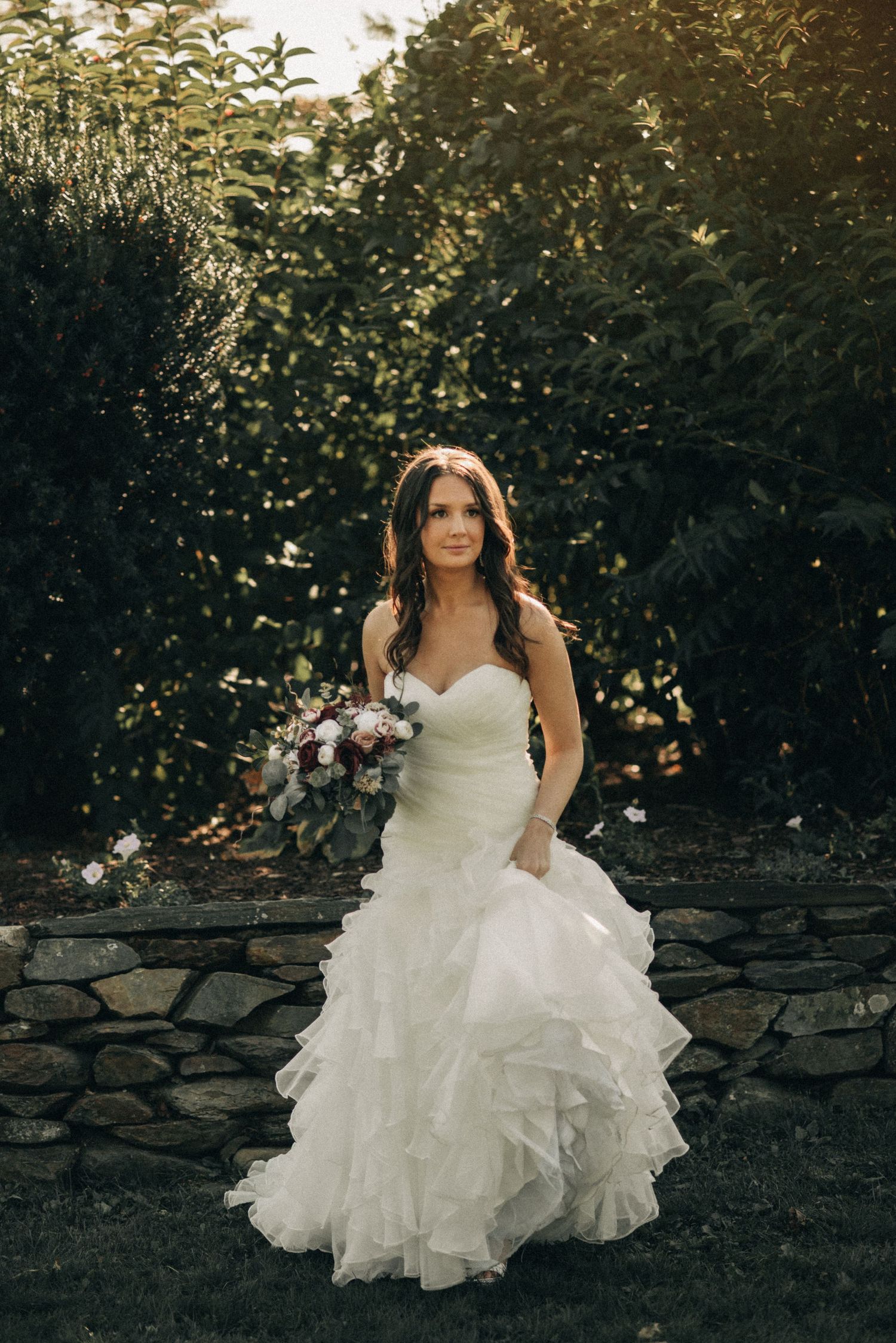 A bride in a ruffled white wedding dress stands in a garden at sunset holding a purple and white bouquet against a stone wall.