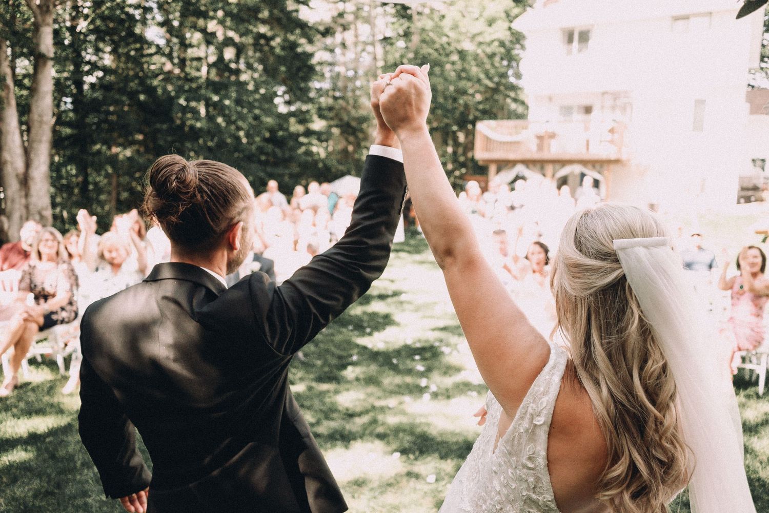A couple raising their arms in celebration during an outdoor wedding ceremony with guests gathered on a lawn.