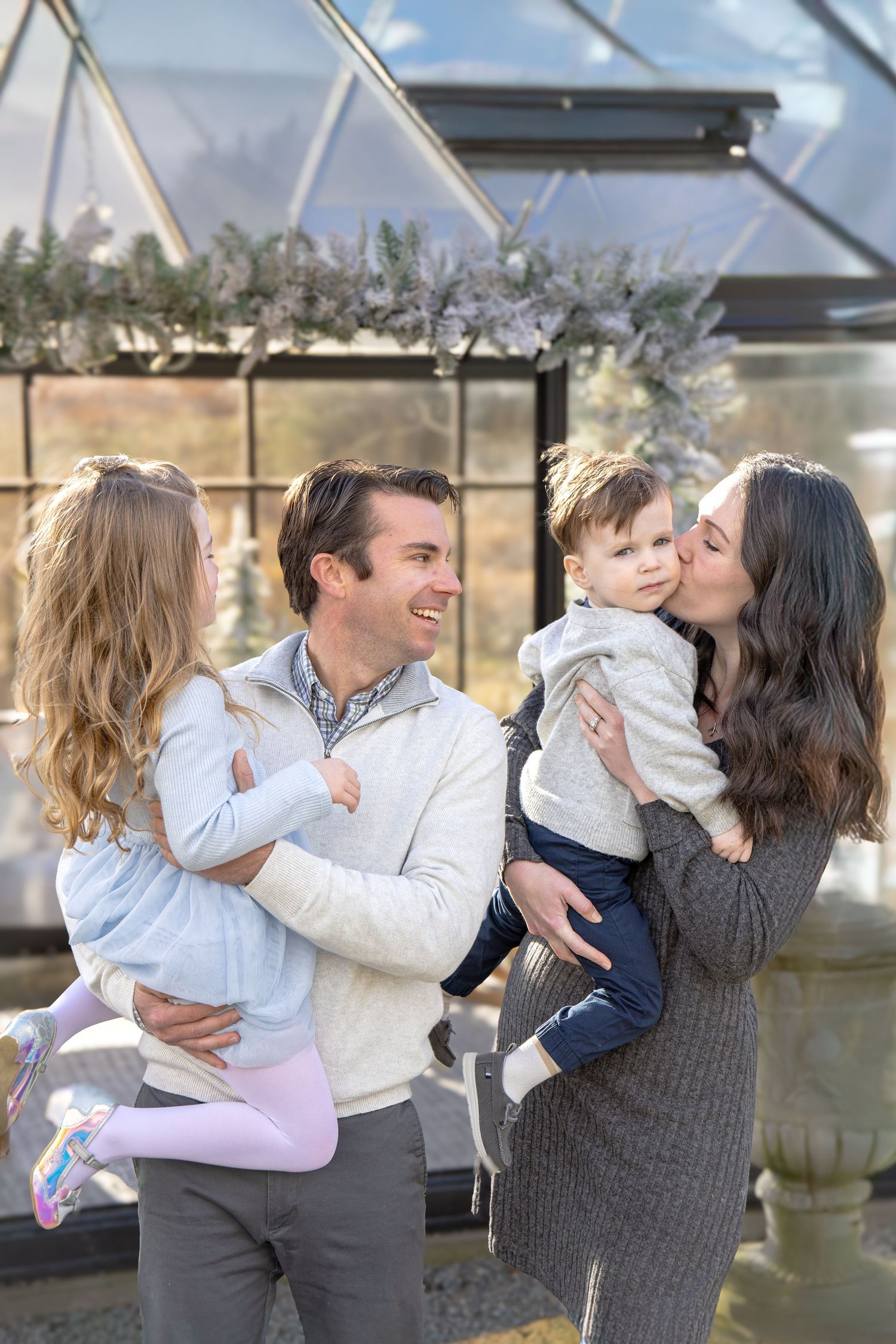 A happy family of four poses outdoors, parents holding young children, smiling and sharing a kiss near a greenhouse.