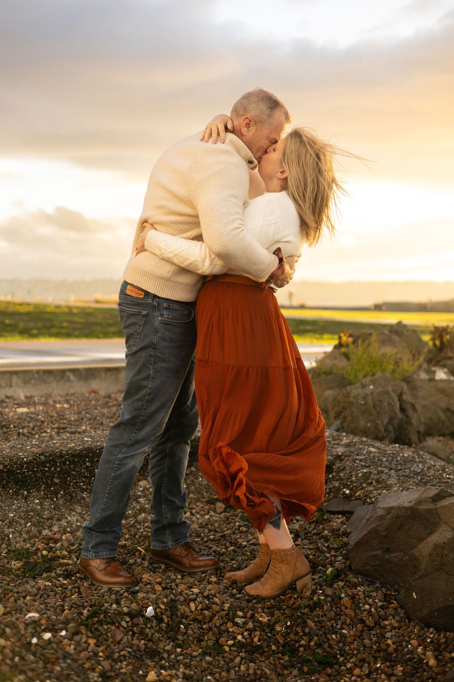 Couple embracing and kissing at sunset on a rocky beach, woman in rust skirt and man in jeans.