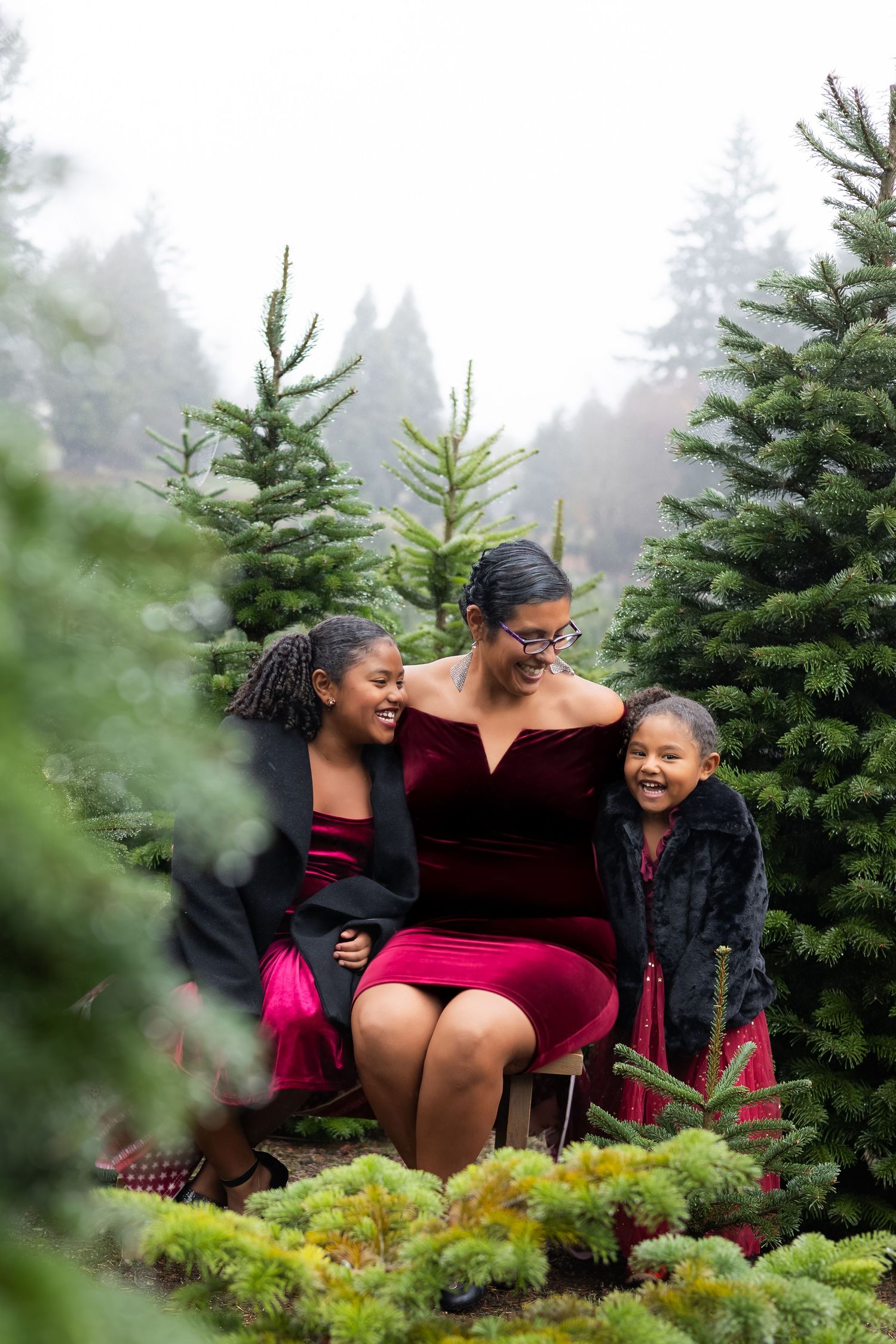 A mother and two daughters laugh together among evergreen trees at a Christmas tree farm, wearing festive red outfits.