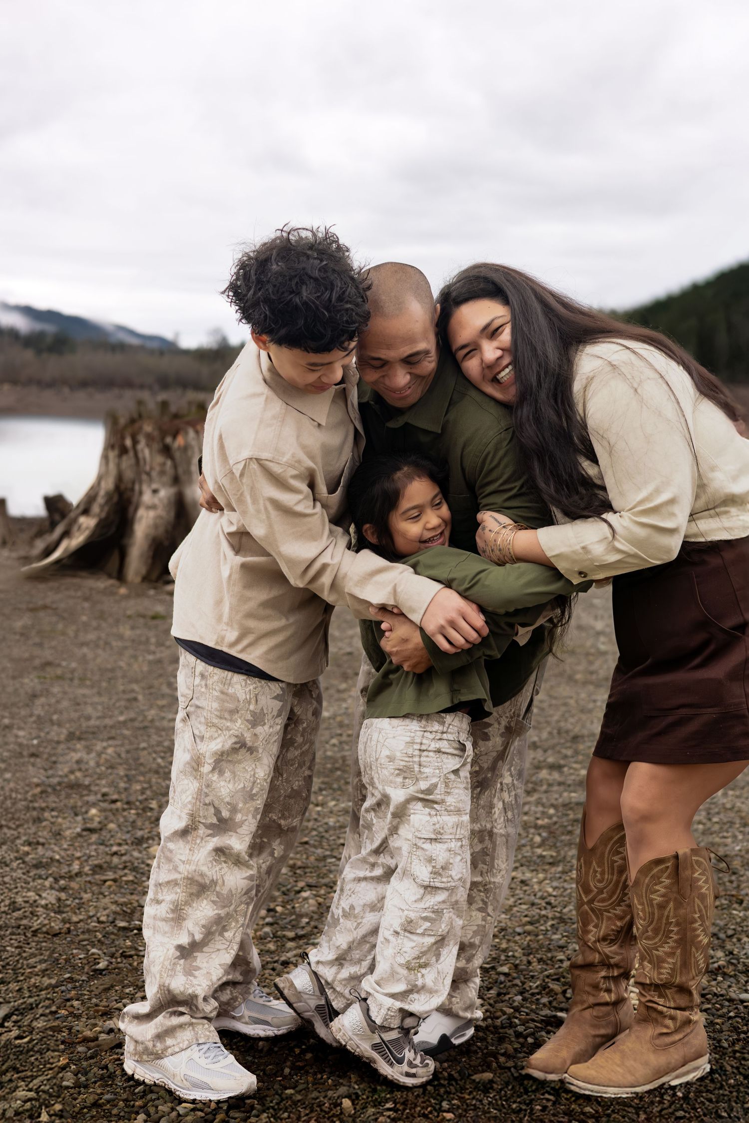 Happy family of four sharing a warm group hug outdoors near a lake with mountains in the background.