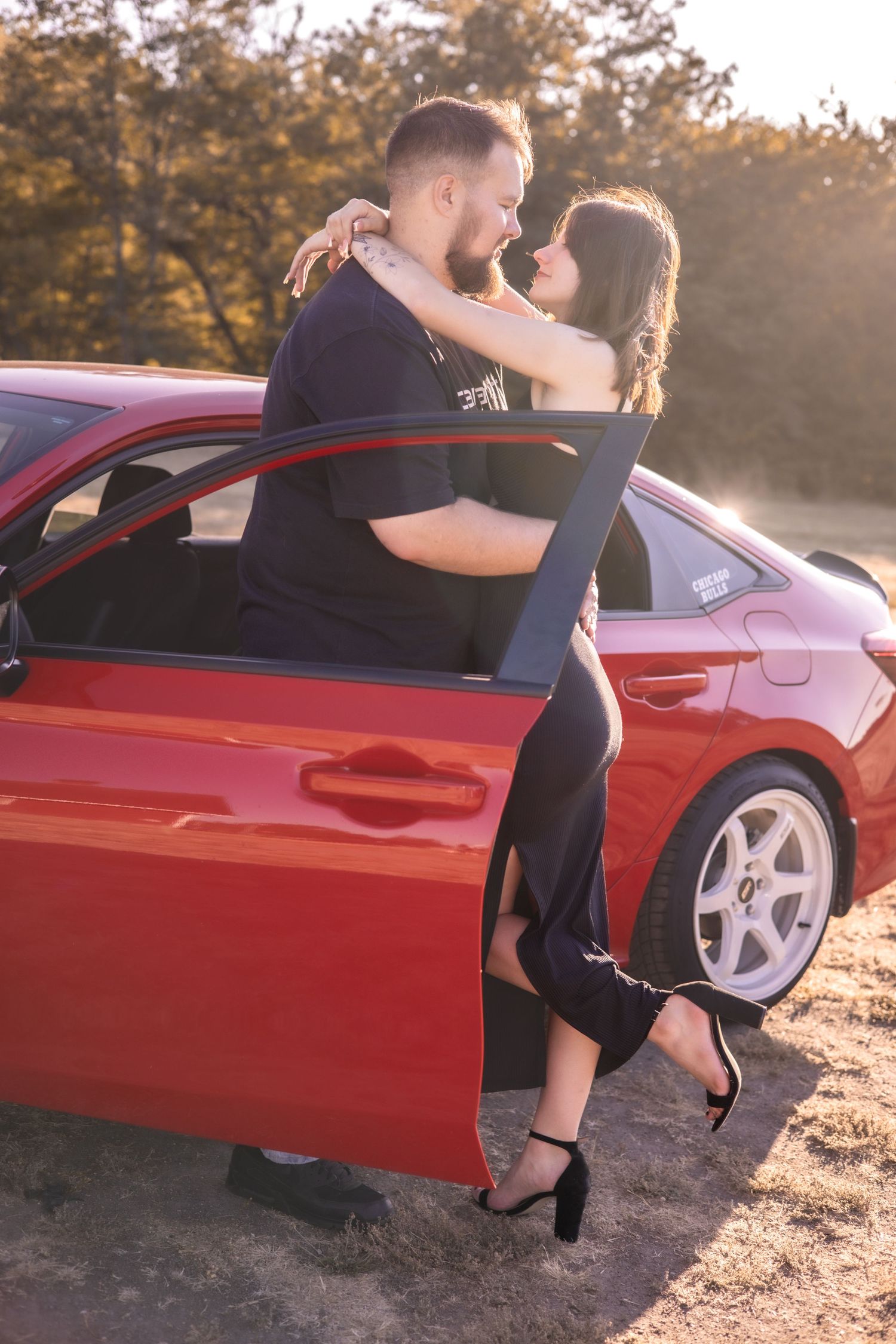 Red sports car serves as backdrop for intimate couple photos at sunset.