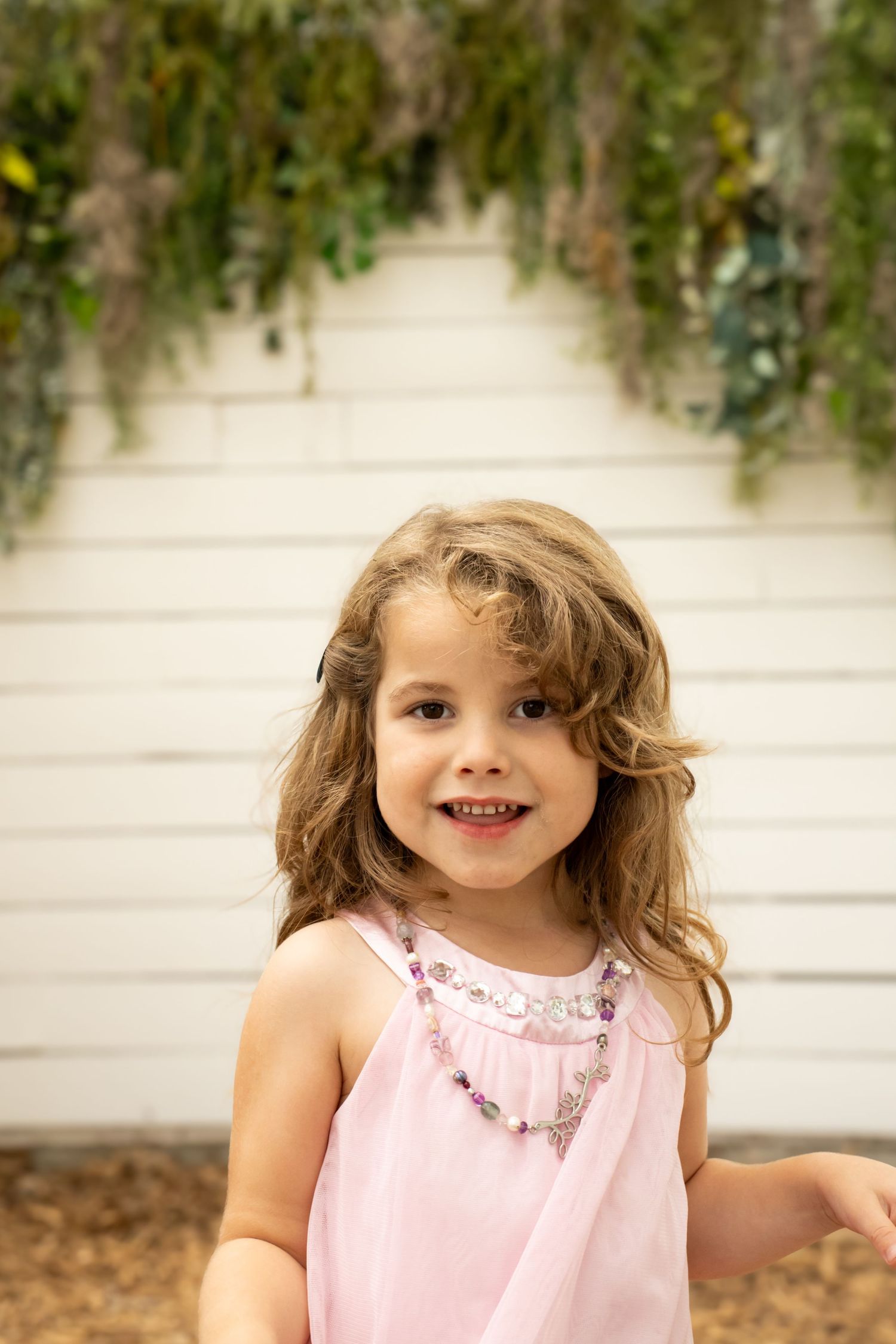 Young girl in pink dress and beaded necklace smiles outdoors near white wall with hanging greenery.