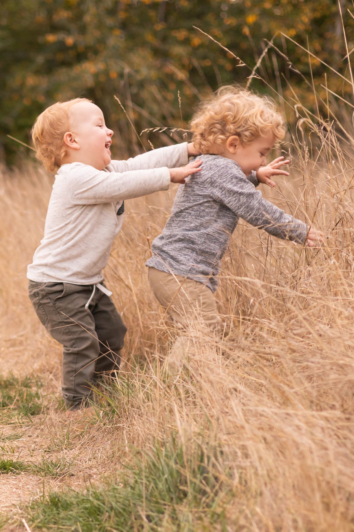 Small child in beige sweater and brown pants plays energetically in tall autumn grass while holding a blanket.