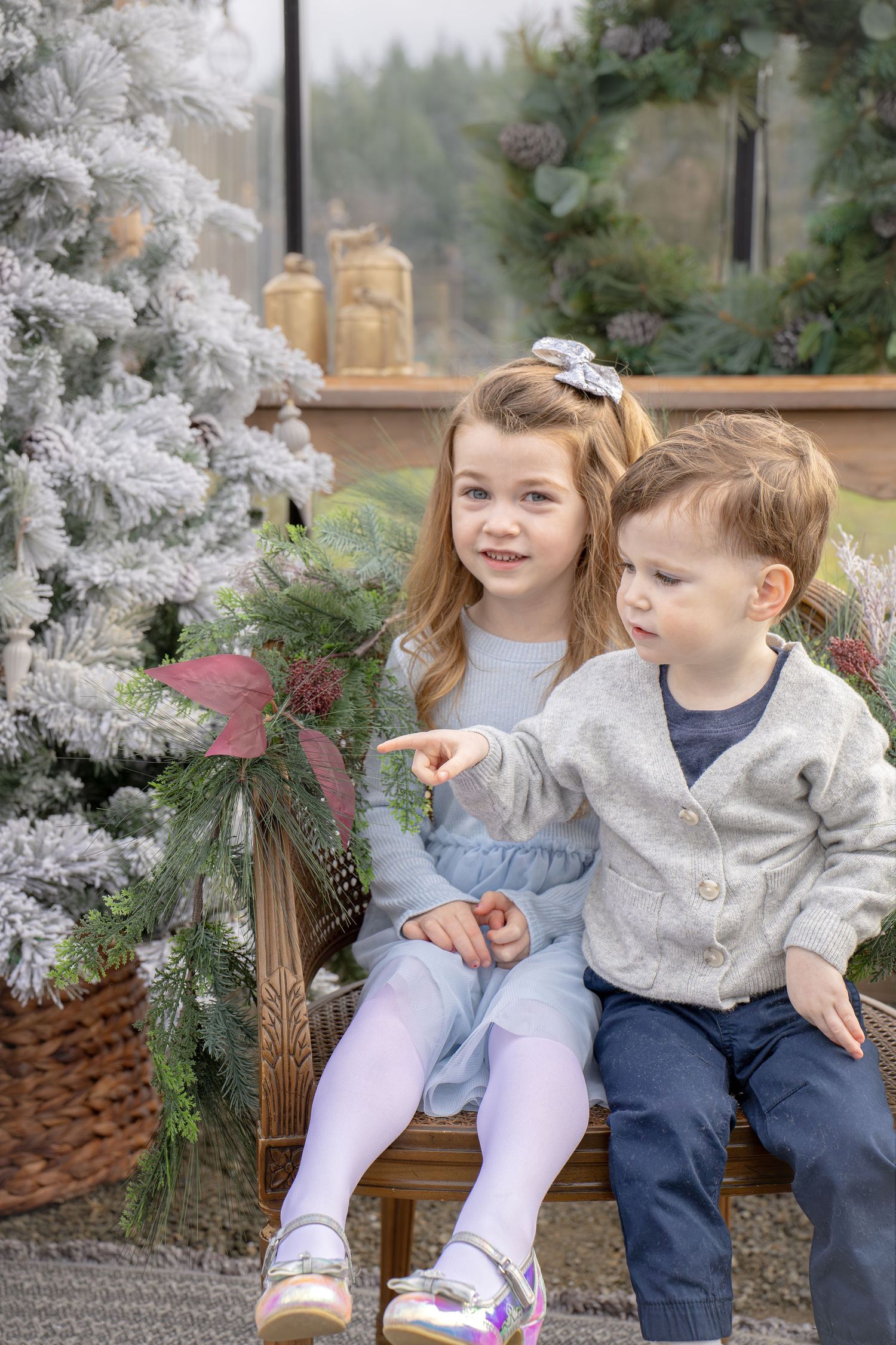 Two children sit together on a bench next to a flocked white Christmas tree with greenery decorations.