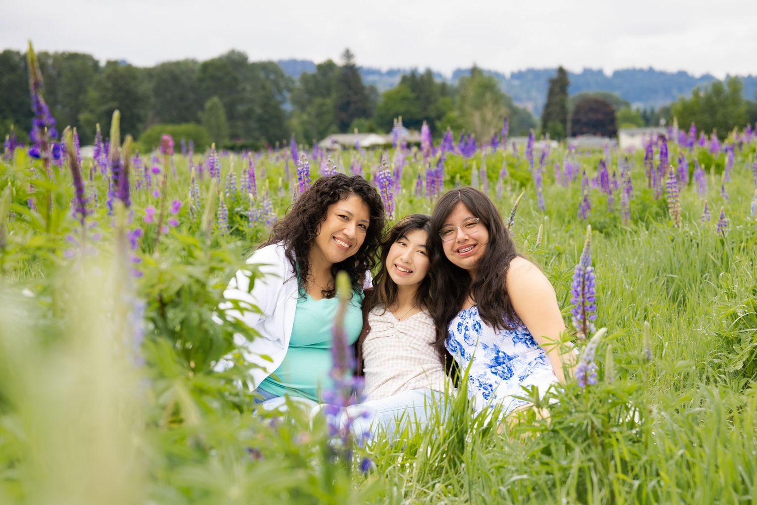 Three women smiling together in a vibrant purple lupine flower field on a cloudy day.