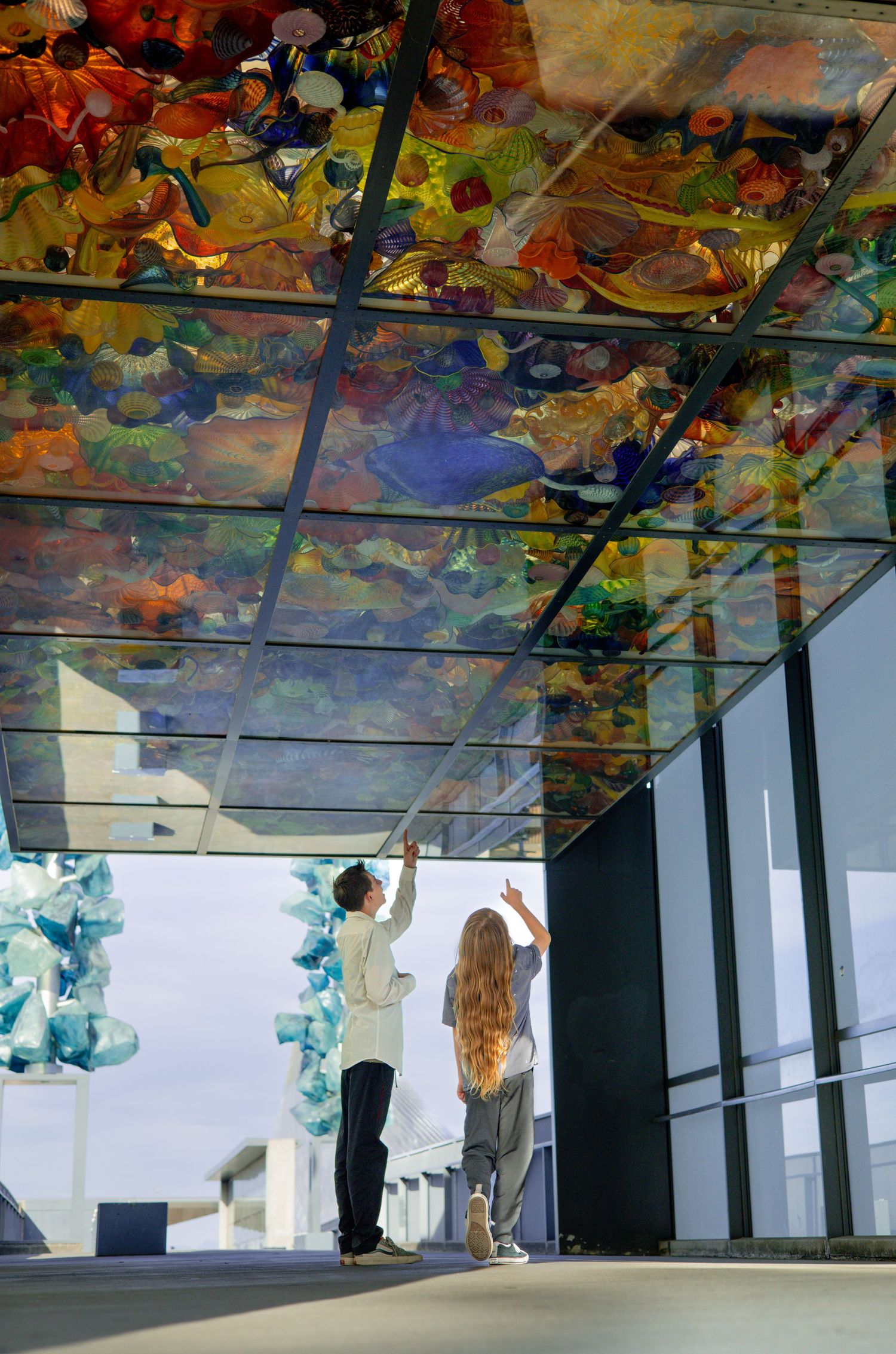 Two people admiring colorful glass art installation on ceiling in modern gallery space.