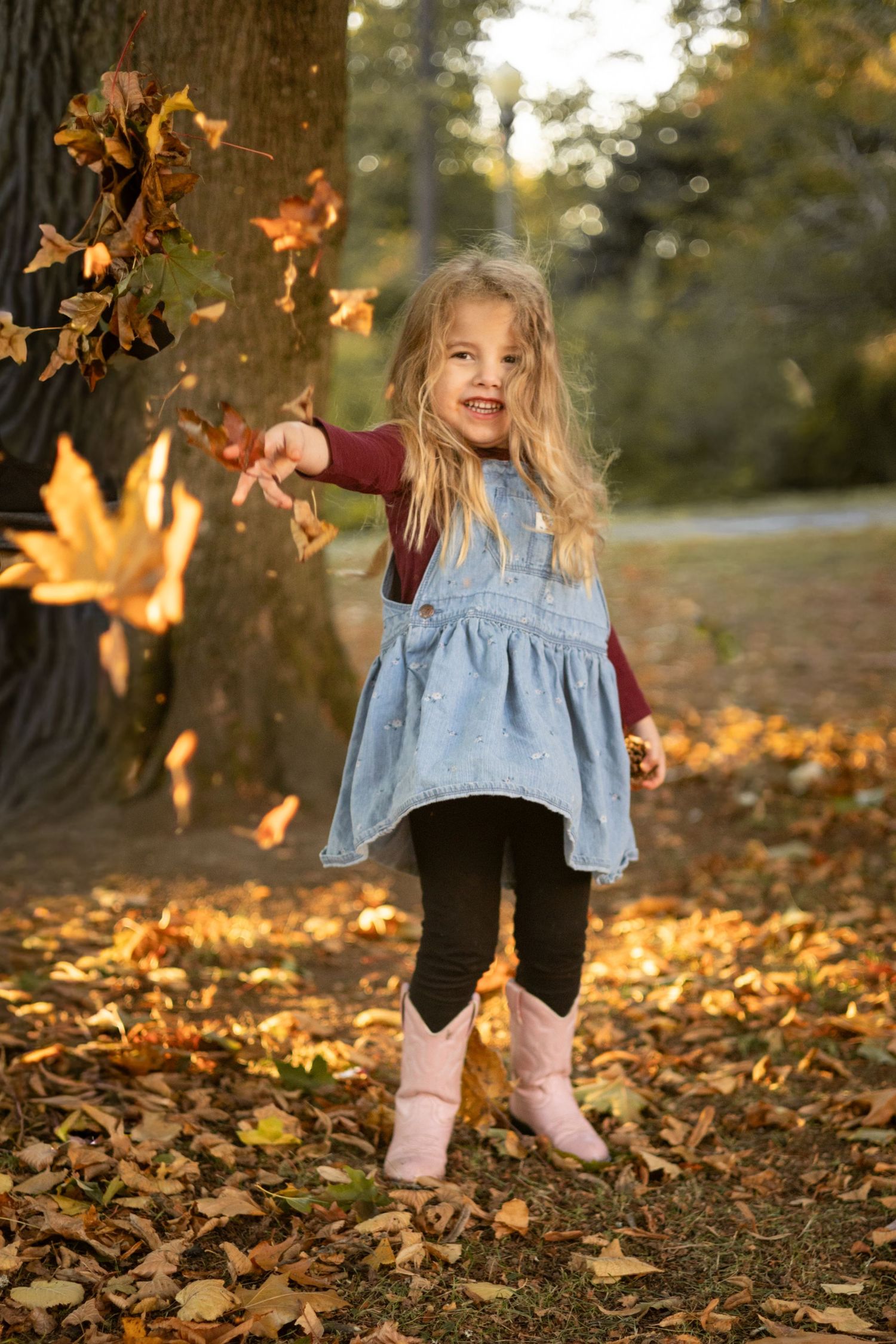 A happy young girl in a denim dress and pink boots tosses autumn leaves in a sunlit park.